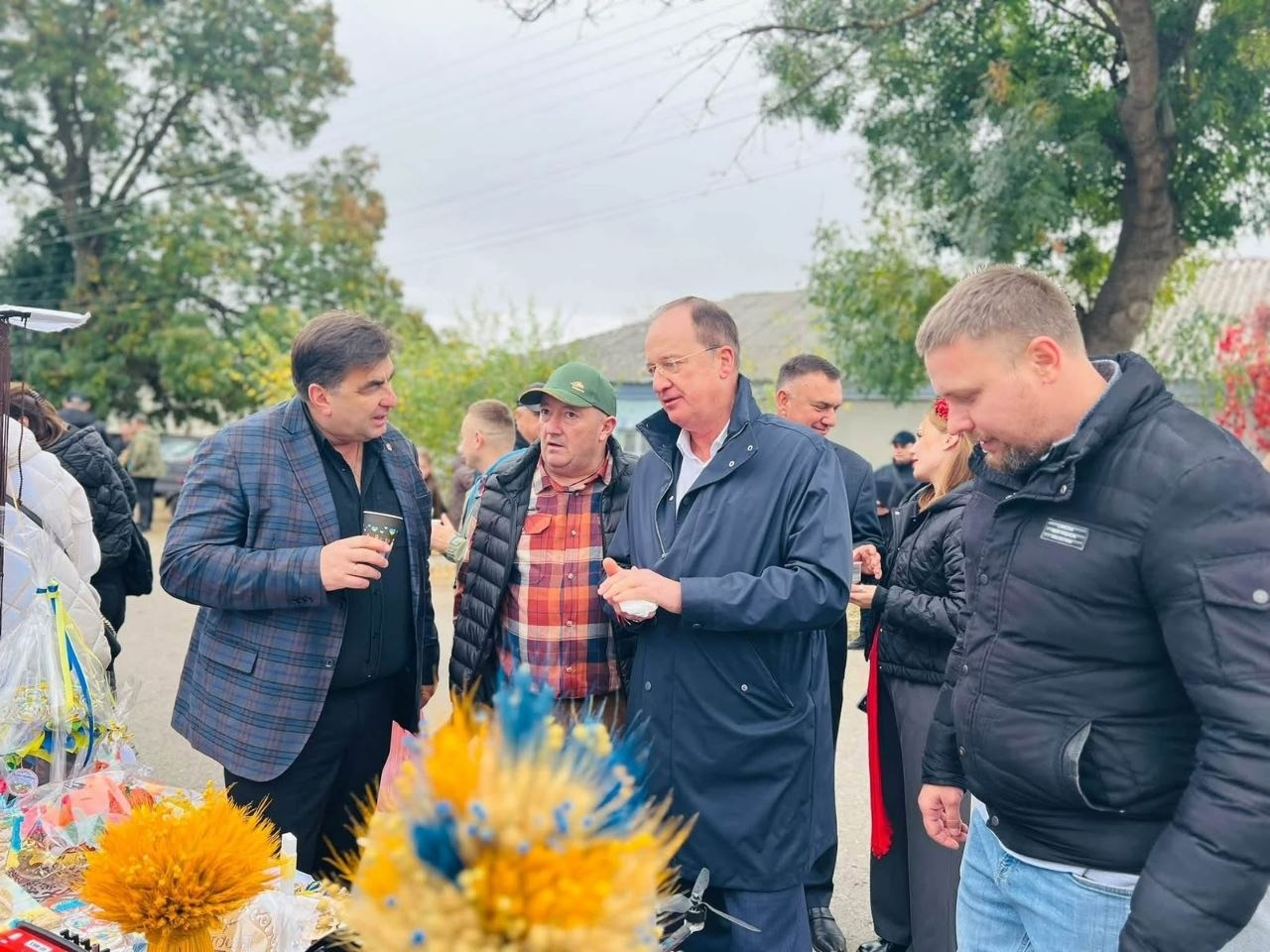 На світлині зафіксовано момент робочого спілкування керівників громад та партнерів під час благодійного ярмарку в селі Курісове. У центрі кадру стоїть чоловік у темно-синьому плащі та окулярах, який веде бесіду з іншими учасниками делегації. По ліву руку від нього (ліворуч у кадрі) перебуває начальник Чулаківської СВА Руслан Русаль у зеленій кепці та картатій сорочці, а поруч із ним — чоловік у синьому картатому піджаку з паперовим стаканчиком у руці. Праву частину кадру займають начальник Бехтерської СВА Олексій Баткалов у чорній куртці (на передньому плані) та чоловік у класичному піджаку. Розмова відбувається просто неба на тлі сільської вулиці з деревами, а на передньому плані ліворуч помітно елементи ярмаркового столу з яскравими патріотичними букетами з сухоцвітів та колосків.