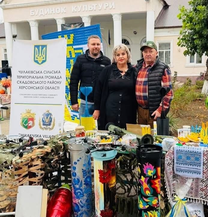 На світлині зафіксовано експозицію Чулаківської громади на благодійному ярмарку в селі Курісове, де на передньому плані представлено військові дрони, маскувальні сітки, розписані тубуси від снарядів, вироби, надруковані на 3D-принтері, та патріотичні сувеніри. На тлі виставки стоять троє представників делегації: праворуч — начальник Чулаківської СВА Руслан Русаль у картатій сорочці та чорній дутій безрукавці, по центру — голова Курісівської громади Наталія Бугайчук у чорній сукні з намистом, а ліворуч — начальник Бехтерської СВА Олексій Баткалов у чорній куртці. З лівого боку кадру встановлено великий білий банер із гербом України та написом «ЧУЛАКІВСЬКА СІЛЬСЬКА ТЕРИТОРІАЛЬНА ГРОМАДА СКАДОВСЬКОГО РАЙОНУ ХЕРСОНСЬКОЇ ОБЛАСТІ», а позаду присутніх височіє будівня з написом «БУДИНОК КУЛЬТУРИ» та велике жовто-блакитне в'язане панно.