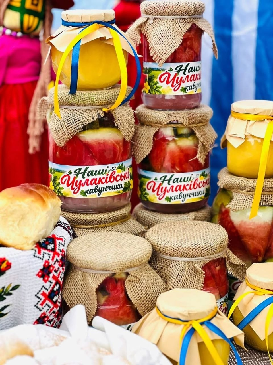На світлині великим планом зображено фірмову продукцію Чулаківської громади — скляні банки з консервованими кавунами, представлені на благодійному ярмарку в селі Курісове. Скляна тарна з червоними скибками кавунів виставлена у кілька ярусів, а три центральні банки мають яскраві білі етикетки з квітковим принтом та написами «Наші Чулаківські кавунчики». Кришки консервації дбайливо задекоровані мішковиною та крафтовим папером, які перев'язані мотузками, а деякі банки додатково оздоблені тонкими синьо-жовтими стрічками. На передньому плані ліворуч лежить свіжа домашня випічка на білій скатертині з традиційним червоно-чорним українським вишитим орнаментом, а на розмитому задньому плані кадру помітно учасників заходу в яскравому національному вбранні та синьо-білу смугасту тканину ярмаркового намету.