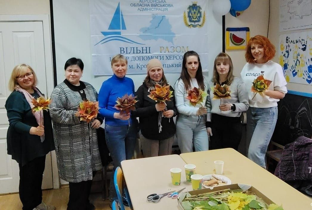 На світлині зображено спільне фото учасниць та майстрині після завершення творчого заняття в координаційному центрі «Вільні разом» у Кропивницькому. Сім жінок вишикувалися в один ряд у приміщенні хабу і з посмішками позують перед об'єктивом, тримаючи в руках виготовлені власноруч оригінальні квіти з різнокольорового осіннього листя. Крайня праворуч стоїть рудоволоса майстриня у білому світшоті та синіх джинсах, яка керувала процесом рукоділля. На передньому плані видно частину навчального столу, де лежать ножиці, картонна коробка з природними матеріалами та тарілка з частуваннями. Задня стіна кімнати прикрашена великим білим банером із офіційним гербом та написом «ХЕРСОНСЬКА ОБЛАСНА ВІЙСЬКОВА АДМІНІСТРАЦІЯ | ВІЛЬНІ РАЗОМ | Центр допомоги херсонцям у м. Кропивницький», а праворуч на стіні помітно патріотичні дитячі малюнки.