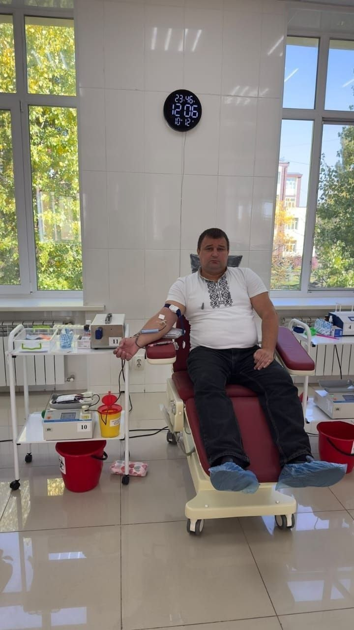 На світлині зображено чоловіка під час здачі крові в медичному закладі в межах кампанії з популяризації донорства від хабу «Вільні разом» у Кропивницькому. Чоловік у білій вишитій футболці-поло, чорних джинсах та бахілах сидить у бордовому донорському кріслі, а з його правої руки здійснюється забір крові. Ліворуч встановлено мобільний медичний столик із вагами-помішувачем для гемоконтейнерів та інструментами під номерами «10» та «11». Маніпуляційна кімната має білі кахельні стіни, де на одній із них висить круглий електронний годинник із червоними цифрами «12 06», а на задньому плані видніються великі вікна з краєвидом на дерева.