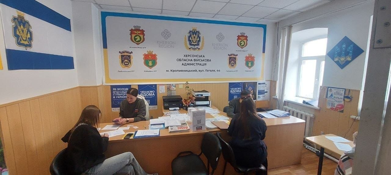На світлині зображено процес надання соціальної допомоги та консультування переселенців із Херсонщини в координаційному центрі «Вільні разом» у Кропивницькому. У робочому кабінеті хабу за двома великими дерев'яними столами співробітники, серед яких мобільна команда БО «100% життя. Кропивницький», ведуть прийом громадян. Ліворуч за столом сидить консультантка, яка оформлює документи для дівчини-переселенки, а праворуч інший фахівець надає роз'яснення жінці, повернутій спиною до об'єктиву. На столах розкладено папки з паперами, канцелярське приладдя, стоїть принтер та інформаційна табличка з QR-кодом. Задня стіна кімнати задекорована великим брендованим жовто-блакитним банером «ХЕРСОНСЬКА ОБЛАСНА ВІЙСЬКОВА АДМІНІСТРАЦІЯ | м. Кропивницький, вул. Гоголя, 44» із гербами громад Херсонщини. На бічній стіні ліворуч висить синьо-білий прапор Чулаківської громади, а під меблями наклеєні патріотичні плакати «ЧЕКАЄМО ВДОМА».