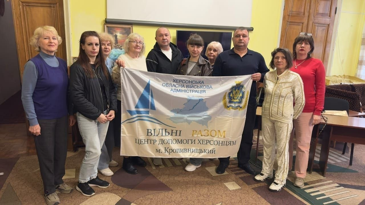 На світлині зображено спільне фото колективу та відвідувачів координаційного центру «Вільні разом» у Кропивницькому. У залі зі світло-жовтими стінами та паркетною підлогою вишикувалася група людей, які тримають перед собою великий білий офіційний банер. На банері нанесено написи: «ХЕРСОНСЬКА ОБЛАСНА ВІЙСЬКОВА АДМІНІСТРАЦІЯ», «ВІЛЬНІ РАЗОМ | ЦЕНТР ДОПОМОГИ ХЕРСОНЦЯМ | м. Кропивницький», а також зображено контурну карту Херсонської області, синій вітрильник та офіційний герб Херсонської області. По центру прапор тримають чоловік у чорній куртці-худі та чоловік у темно-синій футболці-поло, а навколо них стоять жінки у повсякденному одязі (зокрема дівчина у світлих джинсах ліворуч, жінка у світлому костюмі та жінка у яскраво-червоній кофті праворуч). На задньому плані помітно великі дерев'яні міжкімнатні двері, телевізійний екран, невелику картину з морським пейзажем та офісні столи зі стільцями.