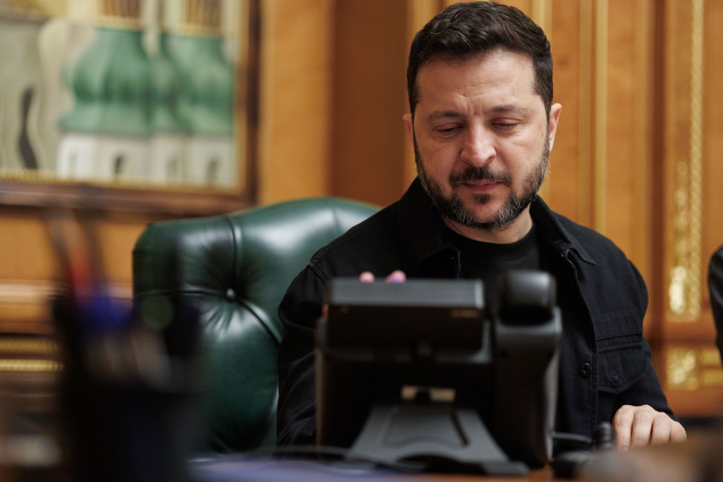 На світлині зображено Президента України Володимира Зеленського під час телефонної розмови у своєму робочому кабінеті. Глава держави сидить у великому шкіряному кріслі темно-зеленого кольору, одягнений у чорну сорочку поверх чорної футболки, і зосереджено дивиться на великий чорний стаціонарний телефон спецзв'язку, що стоїть перед ним на столі. На розмитому задньому плані помітно дерев'яне оздоблення стін кабінету, позолочені елементи інтер'єру та край картини у рамі, а на передньому плані ліворуч видніється підставка для канцелярії.