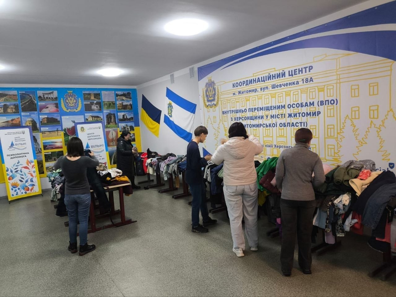 На світлині зображено процес видачі гуманітарної допомоги внутрішньо переміщеним особам із Херсонщини в координаційному центрі «Вільні разом» у місті Житомир. У просторій залі хабу вздовж стін розставлені столи, на яких розкладено велику кількість вживаного сезонного одягу, де відвідувачі самостійно підбирають необхідні речі. На правій стіні закріплено великий біло-жовтий банер із зображенням будівлі та офіційним текстом про підтримку внутрішньо переміщених осіб із Херсонщини у місті Житомир та Житомирській області. Ліворуч на стіні висять Державний Прапор України та синьо-білий прапор Херсонської області, а поруч сформовано великий колаж із фотографій пам'яток Херсонщини під офіційним гербом області. Також у залі встановлено два вертикальних інформаційних ролапи з логотипами хабу: один із написом «ПСИХОЛОГІЧНА ДОПОМОГА» та інший брендований ролап «ВІЛЬНІ РАЗОМ».