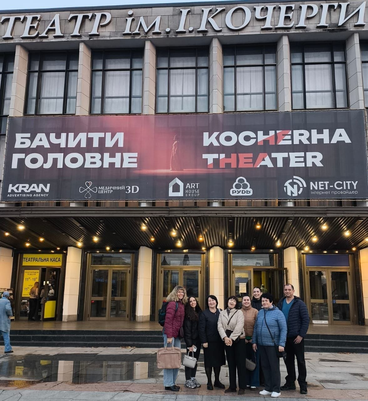На світлині зображено групу внутрішньо переміщених осіб із Херсонщини перед будівлею Житомирського академічного українського музично-драматичного театру імені Івана Кочерги. Сім жінок та один чоловік вишикувалися в один ряд на площі перед центральним входом і з посмішками позують для спільного фото на тлі великих скляних дверей. Фасад театру прикрашений масивним темно-червоним банером із великими написами «БАЧИТИ ГОЛОВНЕ» та «KOCHERHA THEATER», під якими розміщено логотипи офіційних спонсорів та партнерів. Зліва внизу біля вхідної групи помітно жовте віконце з написом «ТЕАТРАЛЬНА КАСА», а на мокрій плитці перед екскурсантами видно дзеркальні відблиски від вечірнього освітлення.