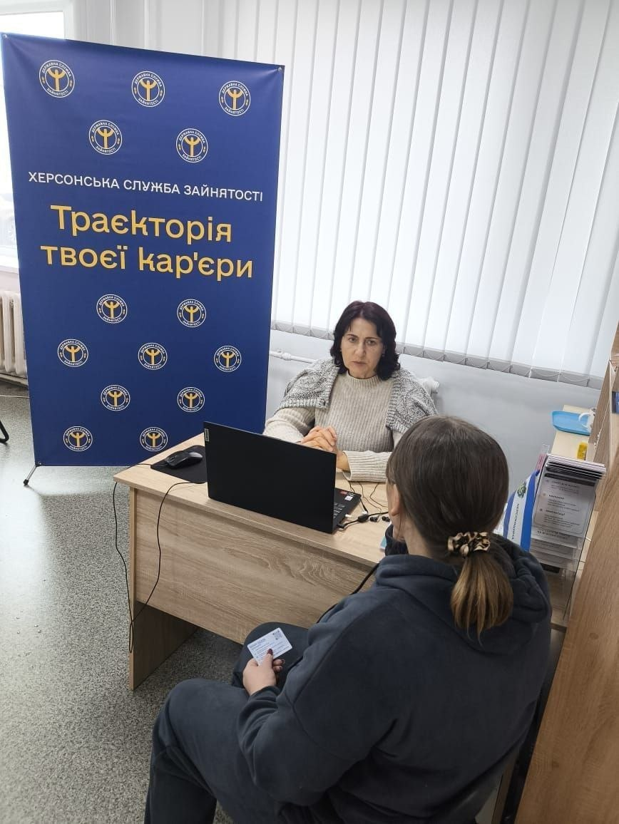 На світлині зафіксовано процес надання індивідуальної консультації з питань працевлаштування в Житомирському координаційному центрі «Вільні разом». За світлим дерев'яним офісним столом сидить фахівчиня Бериславської філії Херсонського обласного центру зайнятості у світлому светрі з накинутою на плечі сірою шаллю. Перед нею стоїть відкритий чорний ноутбук, а навпроти спиною до об'єктиву сидить дівчина-переселенка в темному худі, яка тримає в руці пластикову картку хабу. З лівого боку кадру встановлено великий темно-синій брендований банер Державної служби зайнятості з офіційними золотистими емблемами та написами: «ХЕРСОНСЬКА СЛУЖБА ЗАЙНЯТОСТІ | Траєкторія твоєї кар'єри». Приміщення хабу має білі стіни, вертикальні світлі жалюзі на вікнах та шафу з інформаційними буклетами праворуч.