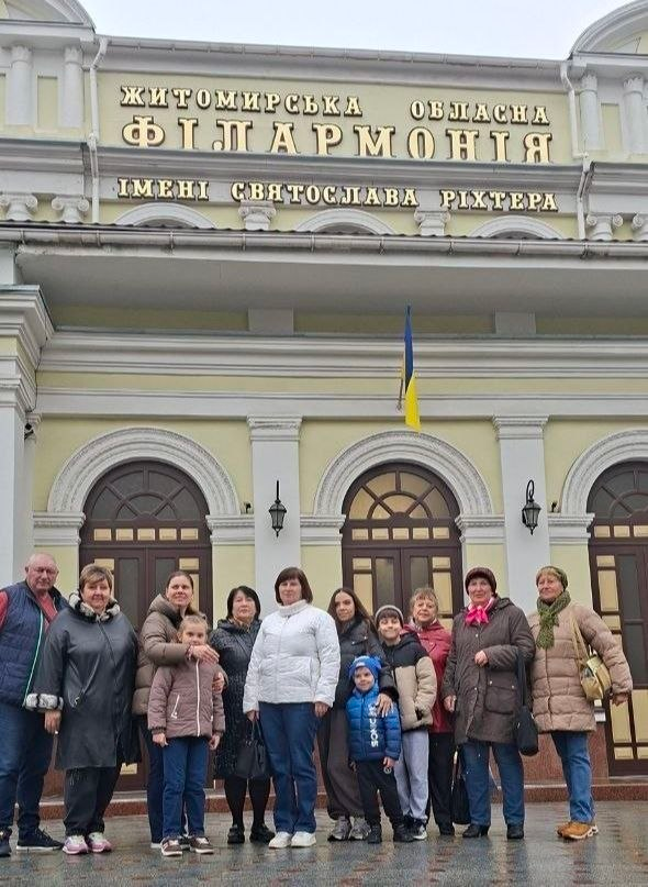 На світлині зафіксовано групу внутрішньо переміщених осіб із Херсонщини перед будівлею Житомирської обласної філармонії імені Святослава Ріхтера, а сам культурний візит організував Житомирський координаційний центр «Вільні разом». Переселенці різного віку та діти вишикувалися в один ряд на площі перед центральним входом на тлі світло-жовтого фасаду з арковими дверима та вікнами, над якими височіє великий золотистий напис: «ЖИТОМИРСЬКА ОБЛАСНА ФІЛАРМОНІЯ ІМЕНІ СВЯТОСЛАВА РІХТЕРА», а по центру над входом закріплено малий Державний Прапор України. Юні гості та дорослі одягнені в теплий повсякденний одяг: осінні пальта, куртки та пуховики різних кольорів. На передньому плані ліворуч стоїть маленька дівчинка в коричневій куртці та синіх джинсах, яку дбайливо обіймає за плечі жінка у коричневій куртці, а в центрі кадру виділяється жінка в яскраво-білій дутій куртці та синіх джинсах. Попереду неї позує маленький хлопчик у синій куртці та шапці, праворуч від нього стоїть ще один хлопчик у бежево-чорній куртці та сірих спортивних штанях, а вже за ними стоїть молода дівчина у темній куртці.