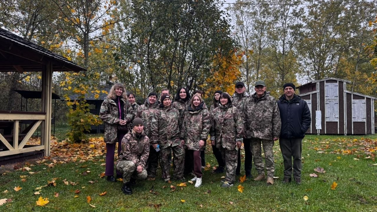 На світлині зафіксовано групу фахівців територіальних громад Херсонщини разом із військовими інструкторами під час проходження Курсу з підготовки громадян до національного спротиву у Житомирі, який організовано за всебічного сприяння місцевого координаційного центру «Вільні разом». Учасники навчання різного віку та статі вишикувалися на великій зеленій галявині, яка рясно вкрита опалим жовто-гарячим осіннім листям. Більшість присутніх одягнені в однакову польову військову камуфляжну амуніцію та штани, а крайній праворуч чоловік стоїть у чорній зимовій куртці та темних тактичних штанях. На передньому плані ліворуч на коліно присіла жінка в камуфляжі та темній шапці. На задньому плані кадру з лівого боку височіє дерев'яна відпочинкова альтанка, з правого боку помітно невеликі дерев'яні господарські будівлі чи кунги, а всю локацію оточує густий осінній ліс із жовто-зеленими кронами дерев.