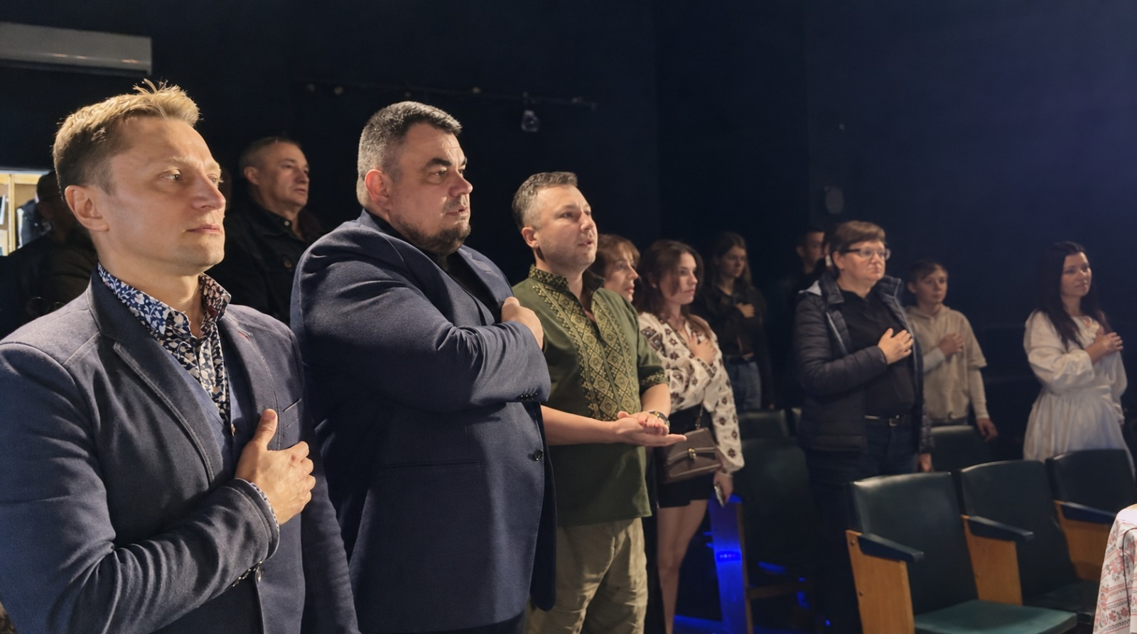 На світлині зафіксовано момент виконання Державного Гімну України під час урочистого заходу з нагоди 210-річчя селища Новотроїцьке, що відбувся у Тернополі. Присутні у залі люди стоять, приклавши праву руку до серця, та зосереджено дивляться вперед. На передньому плані ліворуч стоїть чоловік у синьому піджаку та сорочці з дрібним візерунком, поруч із ним — начальник Новотроїцької СВА Сергій Перетятько у темному класичному костюмі, а далі — чоловік у зеленій вишиванці з традиційним орнаментом. У глибині темного глядацького залу на тлі синіх крісел помітно інших учасників зустрічі, серед яких жінки у вишиваних блузах та повсякденному одязі.