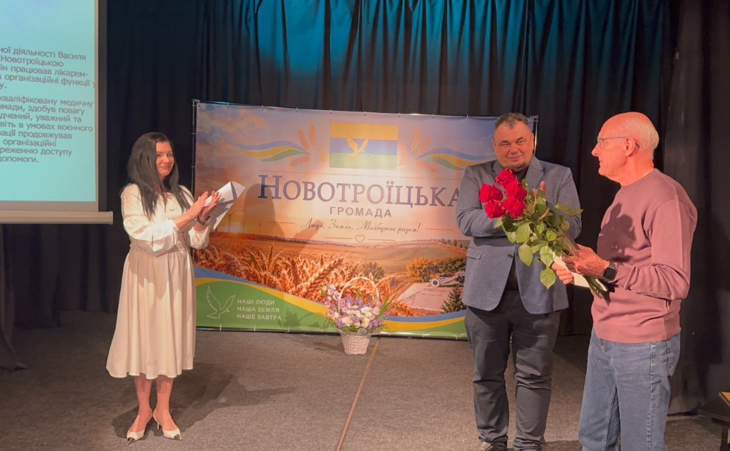На світлині зафіксовано момент урочистого нагородження під час заходу з нагоди 210-річчя селища Новотроїцьке у Тернополі. На сцені на тлі великого офіційного банера з написом «НОВОТРОЇЦЬКА ГРОМАДА | Люди. Земля. Майбутнє разом!» та зображенням пшеничного поля стоять троє людей. По центру начальник Новотроїцької СВА Сергій Перетятько у темному класичному костюмі з посмішкою вручає великий букет червоних троянд літньому сивочолому чоловікові в окулярах та фіолетовому светрі, який стоїть праворуч. Ліворуч від них стоїть дівчина-ведуча у витонченій білій сукні з патріотичною вишивкою, яка радісно аплодує, тримаючи в руках сценарій заходу. Позаду ведучої ліворуч видно частину екрана проєктора з білим текстом, а біля підніжжя банера на підлозі виставлено кошик із ніжно-фіолетовими квітами.