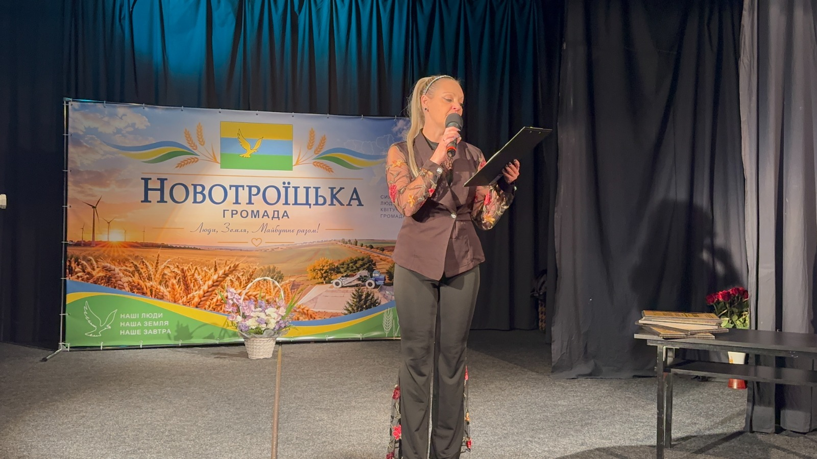 На світлині зафіксовано момент виступу ведучої під час урочистого заходу з нагоди 210-річчя селища Новотроїцьке у Тернополі. Світловолоса ведуча у темній жилетці, чорних штанах та блузі з прозорими квітковими рукавами стоїть праворуч на сцені, тримаючи в лівій руці планшет зі сценарієм, а в правій — мікрофон. Ліворуч за нею розміщено великий офіційний банер із написом «НОВОТРОЇЦЬКА ГРОМАДА | Люди. Земля. Майбутнє разом!» та кошиком квітів біля підніжжя. У правій частині сцени на передньому плані видно темний столик, на якому лежать книги чи папки, а поруч у вазі стоїть невеликий букет червоних троянд.