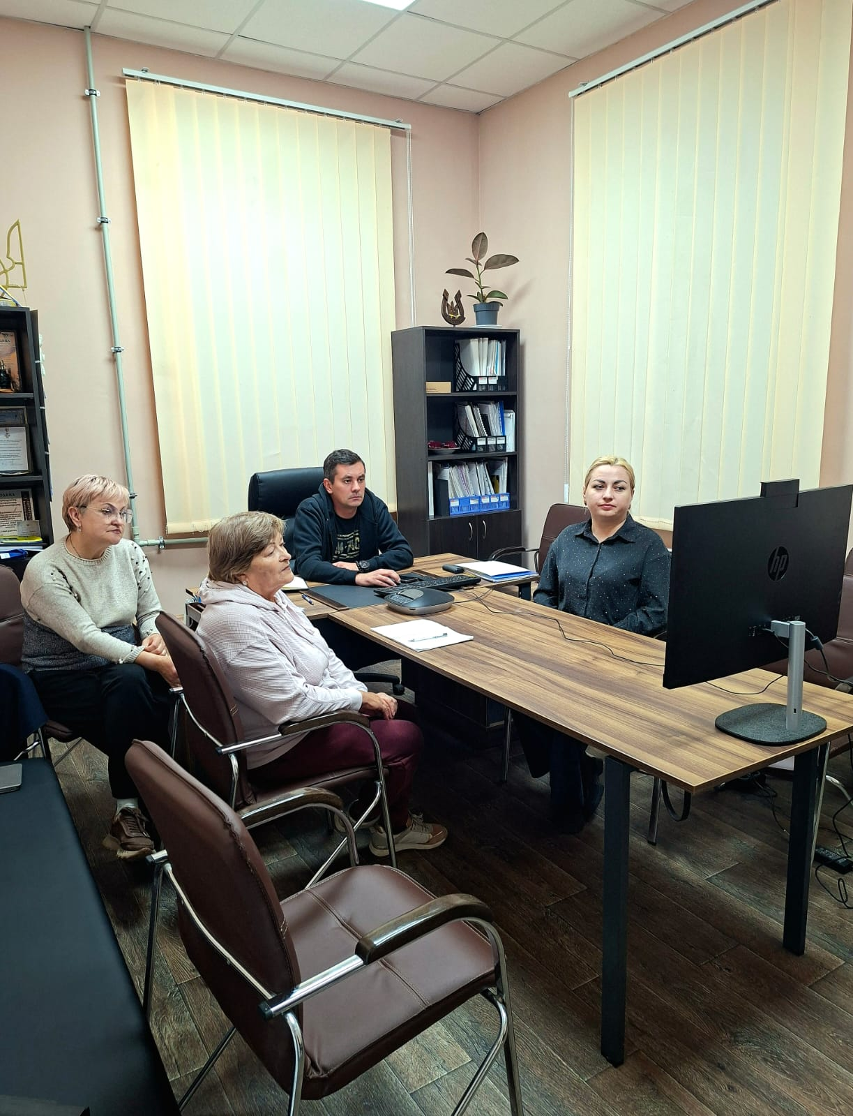 На світлині зафіксовано робочий кабінет під час онлайн-зустрічі представників Високопільської громади з німецькими партнерами. За великим кутовим дерев'яним столом сидять учасники діалогу, які зосереджено дивляться на чорний монітор HP, розташований праворуч. У центрі за клавіатурою перебуває начальник Високопільської СВА Костянтин Стародумов у чорній спортивній кофті, по ліву руку від нього сидить жінка в темній блузі, а ліворуч на офіційних стільцях з коричневою шкіряною оббивкою розмістилися дві жінки старшого віку у світлій кофті та рожевому худі. Кімната має світло-рожеві стіни та підлогу під темне дерево, великі вікна завішені вертикальними тканинними жалюзі бежевого кольору, а на задньому плані між ними височіє чорна офісна шафа з теками для документів та невеликим вазоном на верхній полиці.
