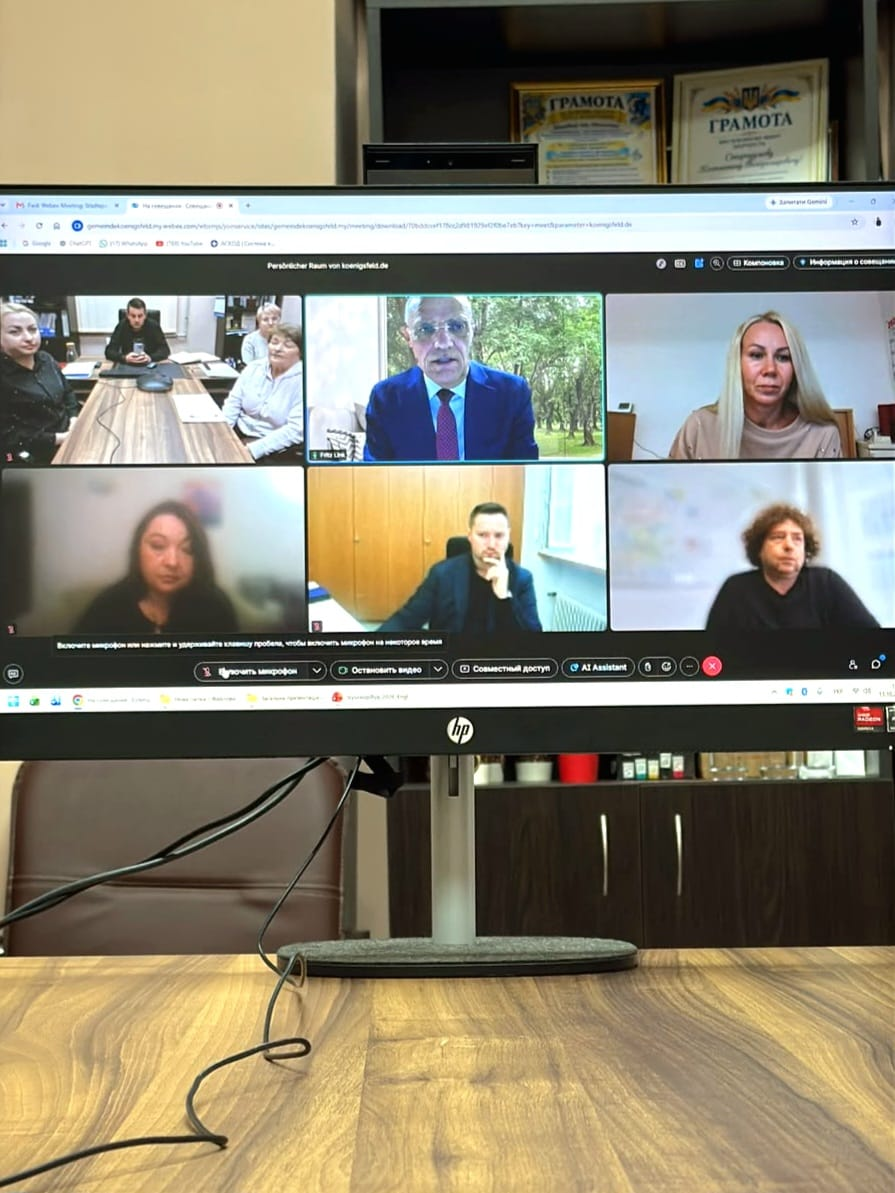 На світлині зафіксовано екран монітора з інтерфейсом онлайн-зустрічі у додатку Cisco Webex, на якому транслюються шість вікон учасників міжнародного діалогу. У верхньому ряду по центру відображається бургомістр громади Кенігсфельд-ім-Шварцвальд Фріц Лінк в окулярах, класичному костюмі та краватці на тлі вікна з парковими деревами, ліворуч у віконці видно кабінет Високопільської СВА з начальником Костянтином Стародумовим та колегами, а праворуч — блондинка у світлій кофті. У нижньому ряду представлені ще троє учасників відеоконференції, серед яких чоловік по центру в чорному піджаці та темній сорочці, який задумливо тримає руку біля обличчя, а також користувачі ліворуч і праворуч на розмитому тлі приміщень. Сам монітор встановлено на світлому дерев'яному столі.