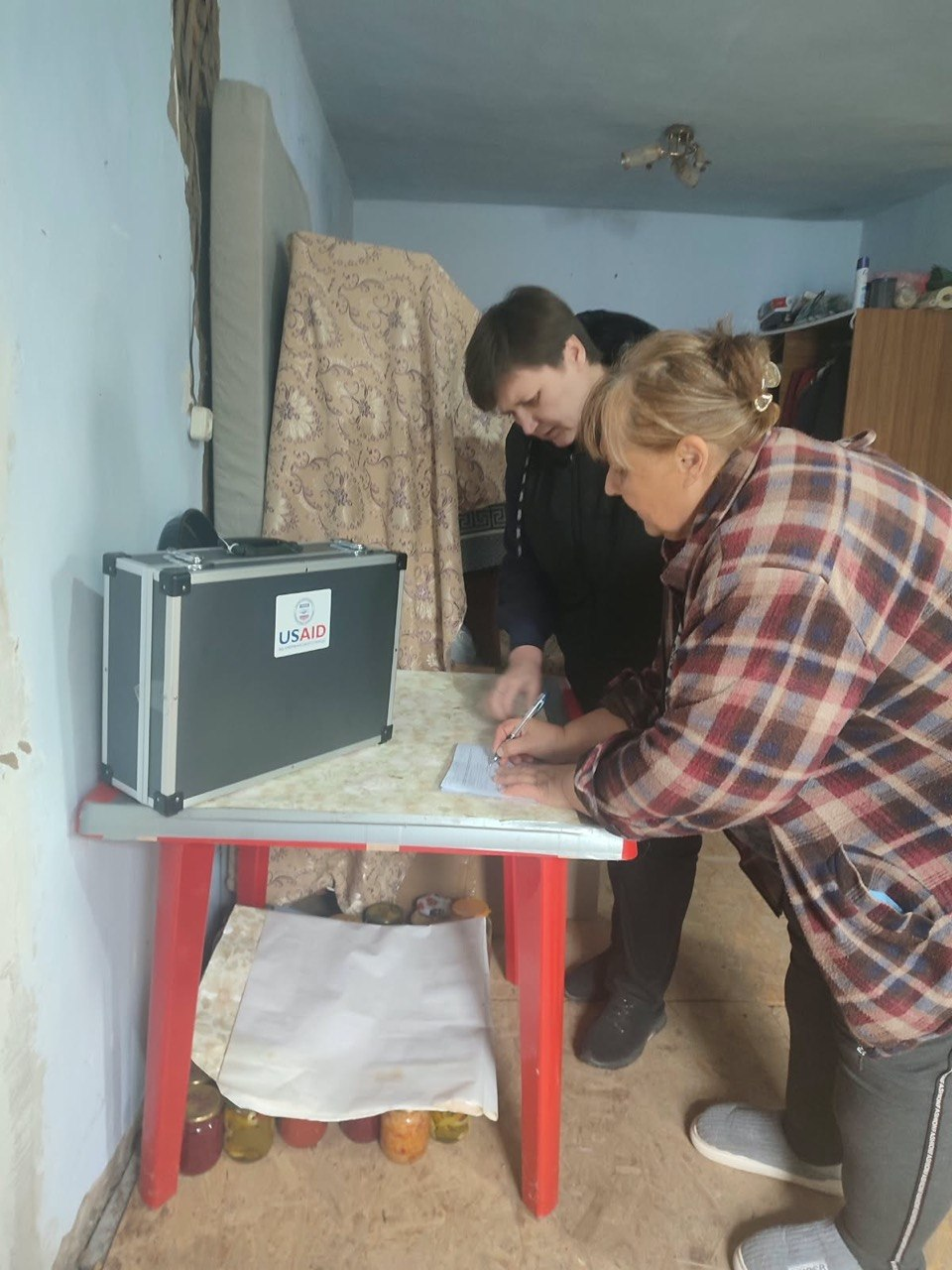 На світлині зафіксовано процес надання виїзних адміністративних послуг жителям Борозенської громади безпосередньо за місцем їхнього проживання. Біля невеликого столу з червоними ніжками стоять дві жінки: місцева жителька у картатій куртці та домашніх капцях зосереджено заповнює або підписує документи, а поруч із нею перебуває адміністратор ЦНАП сільської ради у чорному дутому жилеті, яка координує процес. На столі встановлено велику захищену темно-сіру валізу «мобільного офісу» з офіційним білим логотипом «USAID».