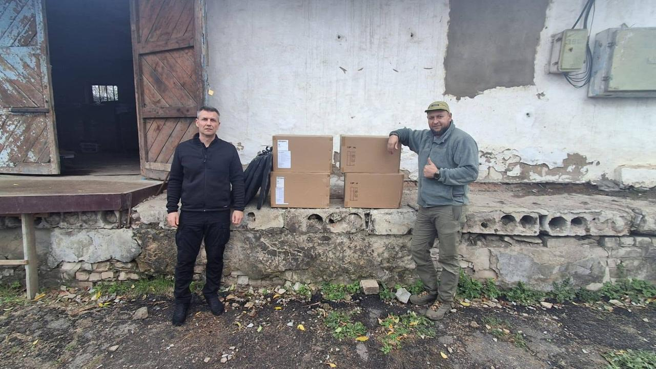 На світлині зафіксовано момент передачі чотирьох зарядних станцій AllPowers для військових 102-ї окремої бригади Сил ТрО. На відкритому майданчику біля старої будівлі зі світлими облупленими стінами та великими дерев'яними воротами стоять двоє чоловіків. Ліворуч на передньому плані позує начальник Бериславської МВА Олександр Алчієв у чорній кофті та чорних тактичних штанях. Праворуч від нього на кам'яному парапеті виставлено чотири великі закриті картонні коробки із зарядними станціями, а поруч із ними — складені сонячні панелі в чорних чохлах. Спершись ліктем на коробки, стоїть військовослужбовець у зеленій флісовій кофті, штанах кольору хакі та кепці, який посміхається і показує великий палець вгору на знак подяки за допомогу.