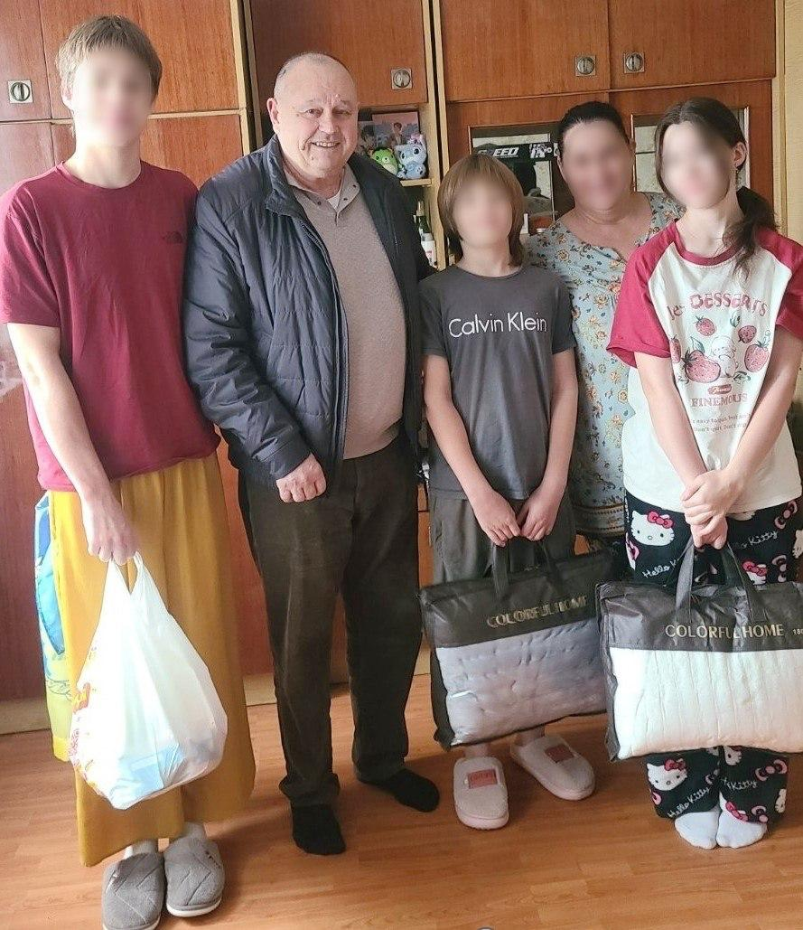 На світлині зафіксовано начальника Таврійської МВА Миколу Різака під час візиту до родини з дітьми, яку повернули з тимчасово окупованих територій і яка наразі мешкає на Рівненщині. У домашній кімнаті з підлогою під світле дерево та дерев'яними шафами на задньому плані для спільного фото вишикувалися п'ятеро людей, при цьому обличчя всіх членів родини (жінки та трьох дітей) на знімку штучно розмиті з міркувань безпеки, а чітким залишається лише обличчя очільника громади. Ліворуч стоїть високий підліток у бордовій футболці та жовтих штанах, який тримає великий білий пластиковий пакет із подарунками. Поруч із ним посміхається очільник громади Микола Різак у темній дутій куртці, сірій кофті-поло та коричневих штанах. Праворуч стоять ще двоє дітей молодшого віку та жінка: хлопчик у сірій футболці бренду «Calvin Klein» та дівчинка у біло-червоній футболці з малюнками солодощів та написом «DESSERTS», які тримають у руках дві великі фірмові прозорі сумки-упаковки з текстильними виробами «COLORFUL HOME». Діти та дорослі взуті у затишні домашні капці, а на полицях шафи на задньому плані помітно м'які іграшки.