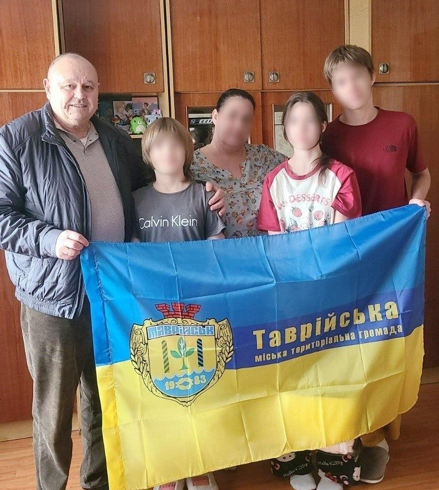 На світлині зафіксовано начальника Таврійської МВА Миколу Різака разом із родиною, яка знайшла прихисток у Рівненській області після повернення з тимчасово окупованих територій. У домашній кімнаті з підлогою під світле дерево та дерев'яними шафами на задньому плані присутні тримають перед собою великий синьо-жовтий прапор із написом «Таврійська міська територіальна громада» та офіційним гербом міста Таврійськ із датою «1983». Обличчя жінки та трьох дітей на знімку штучно розмиті з міркувань безпеки, а чітким залишається лише обличчя очільника громади Миколи Різака, який стоїть крайнім ліворуч у темній дутій куртці. Поруч із ним стоять хлопчик у сірій футболці «Calvin Klein», жінка у квітчастій блузі, дівчинка у біло-червоній футболці з написом «DESSERTS» та підліток у бордовій футболці, які підтримують прапор своєї рідної громади.