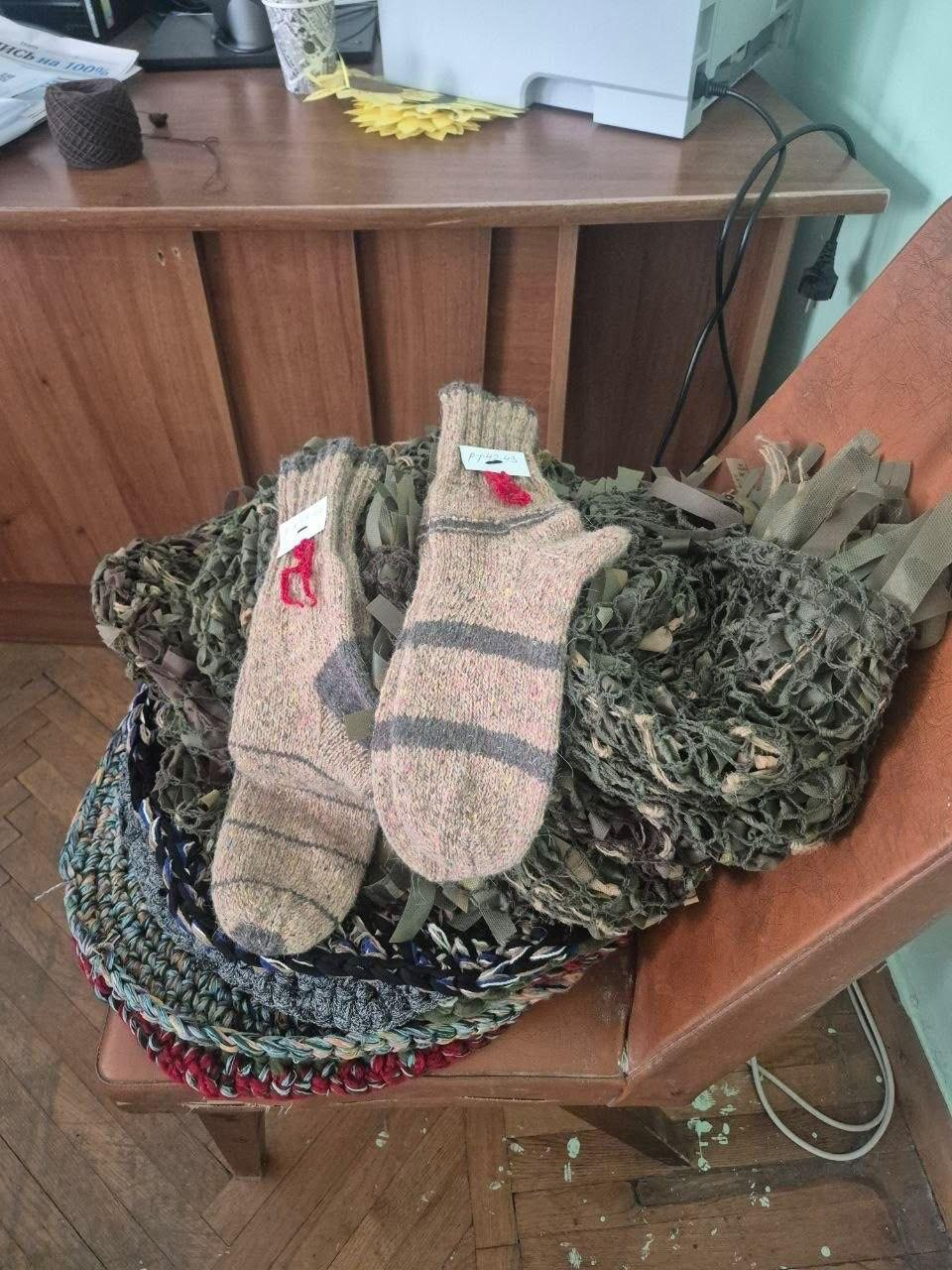 На світлині зафіксовано теплі речі та маскувальні засоби для військових, які переселенки з Херсонщини виготовили в хабі «Вільні разом» у Вінниці. На коричневому шкіряному стільці лежать дві пари плетених вовняних шкарпеток. Під ними складена зелено-коричнева камуфляжна сітка-нашоломник, а нижче видно стопку різнокольорових круглих сидушок. На задньому плані на дерев'яному столі розміщені моток ниток, стаканчик, принтер та друковані матеріали.