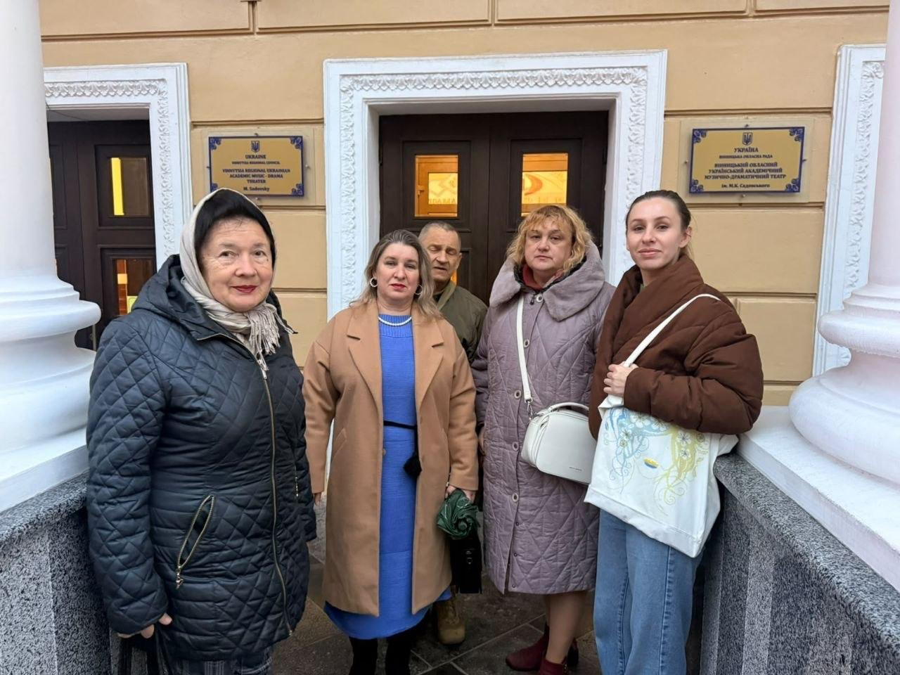 На світлині зафіксовано групу внутрішньо переміщених осіб із Херсонщини перед будівлею Вінницького обласного академічного українського музично-драматичного театру імені Миколи Садовського, а сам культурний візит організував координаційний центр «Вільні разом» у Вінниці. На ганку театру біля масивних світлих колон для спільного фото вишикувалися люди, одягнені в теплий осінній повсякденний одяг: крайня ліворуч стоїть літня жінка у чорній стьобаній куртці та хустці, поруч із нею — жінка у бежевому пальті та синій сукні, а праворуч позують жінка у світло-бузковому пуховику з білою сумочкою та молода дівчина у коричневій куртці з екосумкою, на якій зображено карту України. Позаду між ними на тлі дерев'яних вхідних дверей видно чоловіка, а на світло-жовтій стіні будівлі ліворуч і праворуч закріплено офіційні золотисті інформаційні таблички з назвами театру українською та англійською мовами.