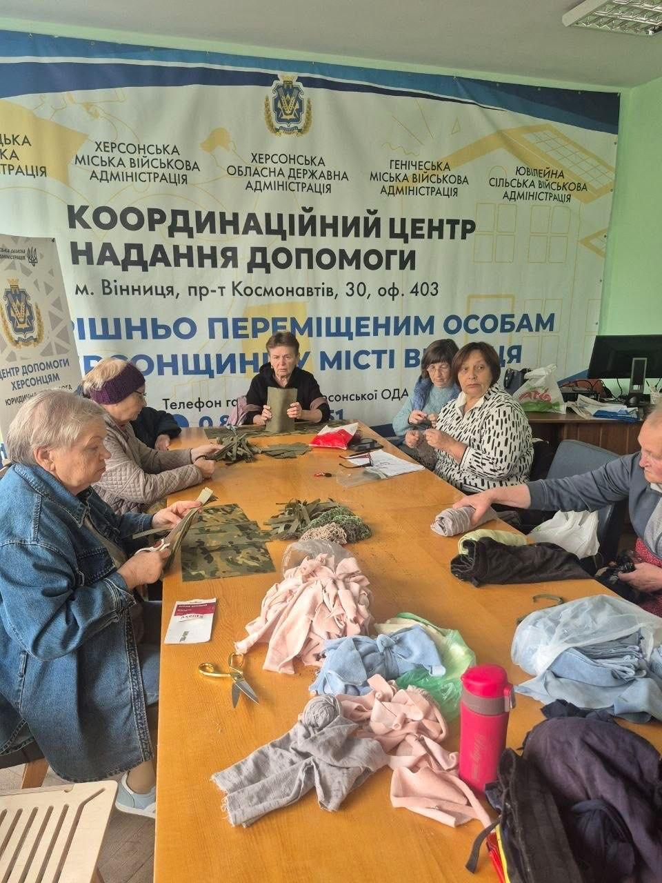 На світлині зафіксовано процес виготовлення маскувальних нашоломників та підготовки речей для українських Захисників і Захисниць переселенками з Херсонщини в координаційному центрі «Вільні разом» у Вінниці. За довгим дерев'яним столом сидять семеро жінок літнього віку, які зосереджено працюють із матеріалами: одні нарізають ножицями смужки камуфляжної тканини мілітарі-відтінків, інші сортують та складають трикотажний і вовняний одяг для формування теплих шкарпеток чи інших текстильних виробів. На столі розкладено клапті тканин, готові плетені елементи, мотки ниток, ножиці, термокружку та інформаційні буклети. За спинами волонтерок височіє великий офіційний біло-зелений банер із гербом України та написами «ХЕРСОНСЬКА МІСЬКА ВІЙСЬКОВА АДМІНІСТРАЦІЯ», «ХЕРСОНСЬКА ОБЛАСНА ДЕРЖАВНА АДМІНІСТРАЦІЯ», «ГЕНІЧЕСЬКА МІСЬКА ВІЙСЬКОВА АДМІНІСТРАЦІЯ», «ЮВІЛЕЙНА СІЛЬСЬКА ВІЙСЬКОВА АДМІНІСТРАЦІЯ», а також великим текстом «КООРДИНАЦІЙНИЙ ЦЕНТР НАДАННЯ ДОПОМОГИ» для підтримки внутрішньо переміщених осіб із Херсонщини у місті Вінниця.