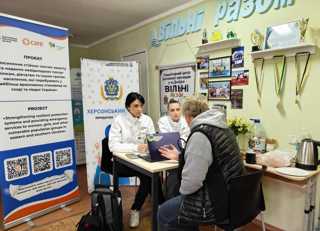 На світлині зафіксовано процес надання індивідуальної юридичної консультації переселенці з Херсонщини від партнерських організацій у координаційному центрі «Вільні разом» у Дніпрі. За невеликим дерев'яним столом сидять дві фахівчині в білих фірмових кофтах із логотипами організації Care, які роз'яснюють правові питання за допомогою відкритого ноутбука. Навпроти них спиною до об'єктиву сидить літня жінка-переселенка в темній стьобаній жилетці та сірій кофті. Зліва в кадрі встановлено великий інформаційний ролап проєкту «Посилення стійких систем захисту та надання невідкладних послуг жінкам, дівчатам та іншим групам» українською та англійською мовами з трьома QR-кодами, а поруч стоїть вертикальний банер із гербом України та написом «ХЕРСОНСЬКИЙ ХАБ ЮРИДИЧНА ДОПОМОГА». Офісне приміщення хабу має світло-жовті стіни, вгорі закріплено об'ємні літери гасла «ВІЛЬНІ РАЗОМ», а на бічній стіні праворуч розміщено полицю з грамотами, спортивними кубками та нагородними медалями на жовто-блакитних стрічках.