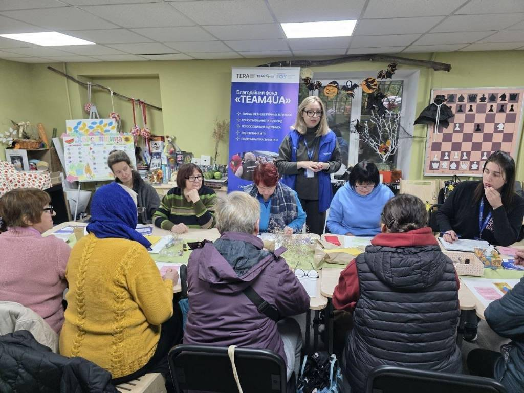 На світлині зафіксовано заняття з безбар’єрного мовлення та арттерапію з виготовлення свічок із вощини від БФ «TEAM4UA» у хабі «Вільні разом» у Дніпрі. За великим столом сидить група літніх жінок-переселенок, які займаються творчістю, а по центру стоїть молода лекторка фонду в окулярах та синій фірмовій жилетці з бейджем. Позаду неї встановлено великий фіолетовий ролап «Благодійний фонд «TEAM4UA», а крайня праворуч за столом сидить інша представниця організації, яка робить записи. Навколо столу, де розкладено матеріали для свічок, розмістилися відвідувачки в осінньому одязі (зокрема жінка у жовтому светрі та синій хустці ліворуч). Оливково-зелені стіни кабінету прикрашені паперовими кажанами, ліворуч стоїть плакат «Херсонщина», а на правій стіні висить демонстраційна шахова дошка.