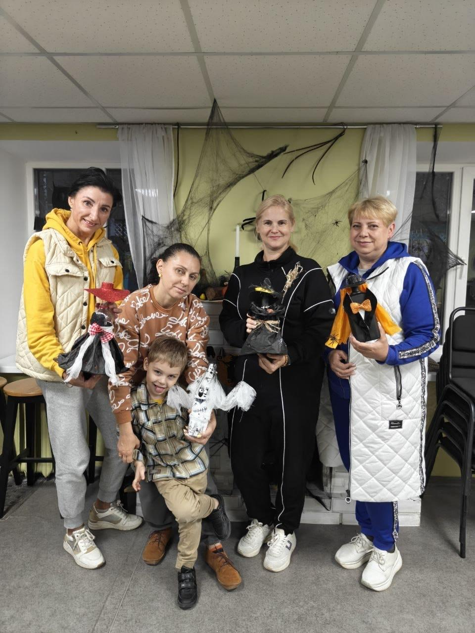 На світлині зафіксовано учасників артстудії «Веселий кавун» із виготовленими декоративними прикрасами до Геловіну в координаційному центрі «Вільні разом» у Дніпрі. Для спільного фото позують чотири жінки та маленький хлопчик, які тримають у руках саморобні тематичні ляльки-мотанки у вигляді відьом у капелюхах та білого привіда. Крайня ліворуч стоїть жінка в жовтому худі та світлому дутому жилеті, поруч із нею присіла жінка в коричневому світшоті з візерунками, яка обіймає хлопчика в картатій сорочці. Праворуч стоять жінка в чорному спортивному костюмі та жінка в синій кофті й довгому білому жилеті. Світло-зелена стіна та декоративний камін на задньому плані кімнати прикрашені густим чорним штучним павутинням із павуками та білими тюлевими фіранками під підвісною стелею «Армстронг».