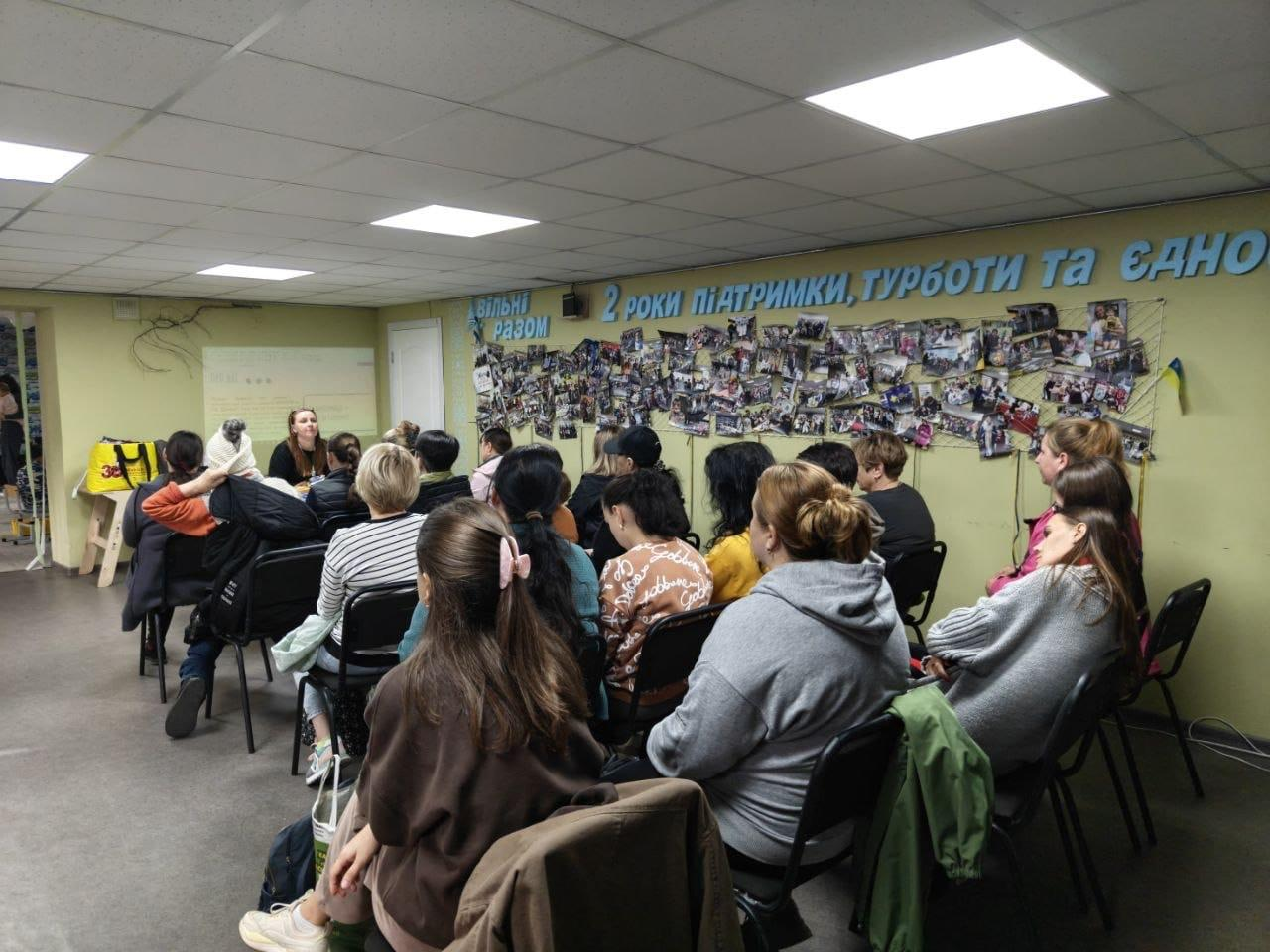 На світлині зафіксовано зустріч для підлітків на тему довіри у відносинах у межах проєкту «Girls Shine» від ГО «Десяте квітня», яка відбулася в координаційному центрі «Вільні разом» у Дніпрі. У просторій залі зі світло-зеленими стінами на стільцях сидить велика група учасниць спиною до об'єктиву, які уважно слухають лекторку, що веде лекцію за столом на задньому плані ліворуч. Уся задня стіна хабу прикрашена масштабним патріотичним фотоколажем із сотень маленьких знімків про життя центру, над якими закріплено великий об'ємний блакитний напис: «2 РОКИ ПІДТРИМКИ, ТУРБОТИ ТА ЄДНОСТІ», а ліворуч на початку цієї інсталяції видніється логотип «ВІЛЬНІ РАЗОМ». Присутні дівчата та жінки одягнені в повсякденний осінній одяг (пальта, кофти, світшоти), на передньому плані ліворуч сидить дівчина з рожевою шпилькою-бантом у волосі, а на стелі горять квадратні вбудовані лампи денного світла.