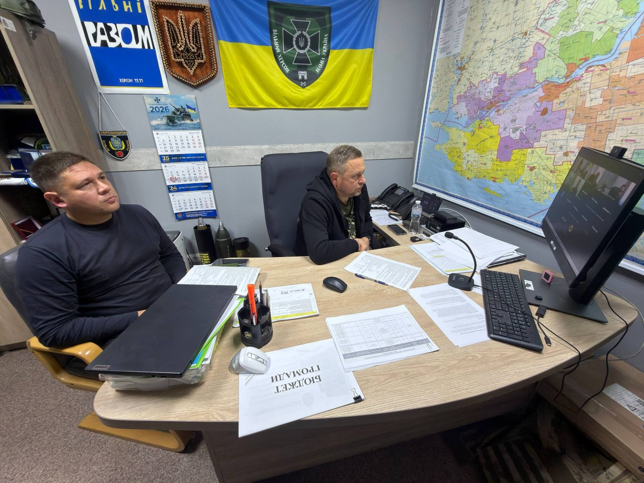 На світлині зафіксовано першого заступника начальника Херсонської ОВА Дмитра Бутрія та директора Департаменту розвитку сільського господарства та зрошення ХОДА Дмитра Юнусова під час онлайн-наради з питань підтримки аграріїв. Посадовці сидять за великим світлим дерев'яним столом у робочому кабінеті й зосереджено дивляться на чорний монітор праворуч, де відображається вікно відеоконференції. Ліворуч у темному світшоті сидить Дмитро Юнусов, а по центру в чорній кофті зафіксований Дмитро Бутрій. На столі перед ними розкладено робочі документи, серед яких папка з написом «БЮДЖЕТ ГРОМАДИ», стоїть відкритий ноутбук Lenovo, комп'ютерна периферія та настільний мікрофон. Стіни кабінету прикрашені Державним Прапором України з військовою символікою, дерев'яним гербом України, емблемою «ВІЛЬНІ РАЗОМ», настінним календарем на 2026 рік, а на правій стіні розвішана велика детальна карта Херсонської області.