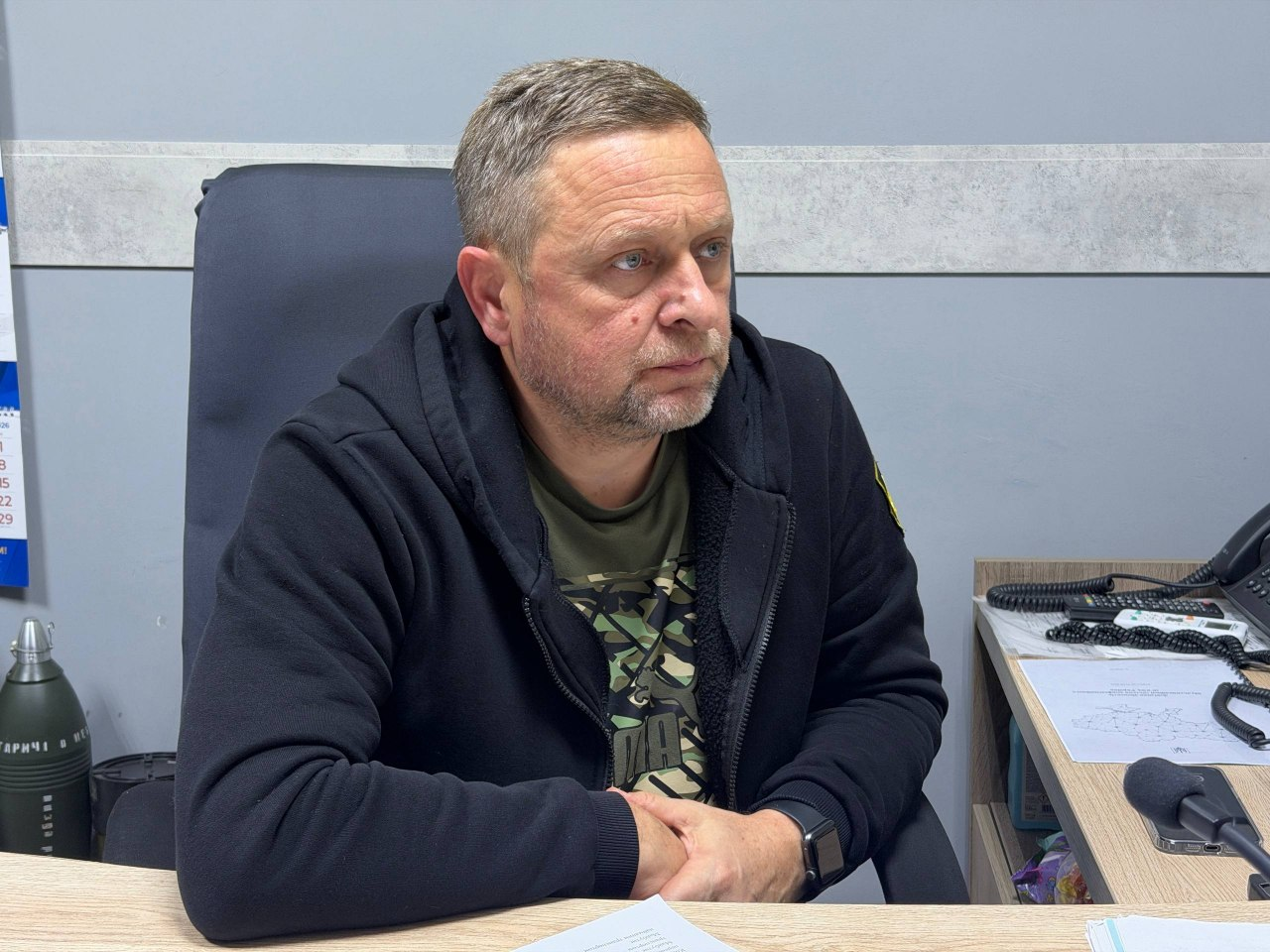 На світлині зафіксовано першого заступника начальника Херсонської ОВА Дмитра Бутрія під час онлайн-наради щодо розробки механізму компенсації логістичних витрат для аграріїв прифронтових територій. Посадовець сидить у великому офісному кріслі з високою чорною спинкою та зосереджено дивиться праворуч перед собою, зчепивши пальці рук на столі. Він одягнений у чорну спортивну кофту із застібкою-блискавкою, під якою помітно футболку кольору хакі з камуфляжним принтом. На його лівій руці закріплено чорний смартгодинник. Робоче місце облаштоване в кабінеті зі світло-сірими стінами, де ліворуч стоїть великий макет артилерійського снаряда з написами та видніється настінний календар. Праворуч на світлому дерев'яному столі лежать робочі документи з роздрукованими картами, встановлено настільний мікрофон на гнучкій ніжці та стаціонарний телефонний апарат чорного кольору.