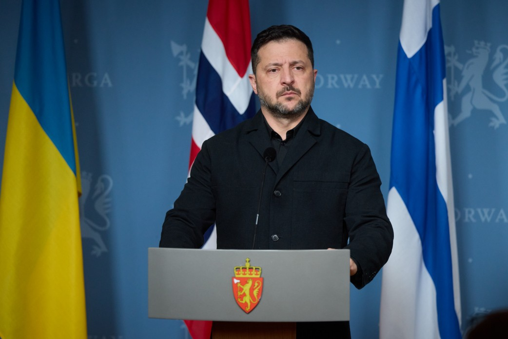 На світлині зафіксовано Президента України Володимира Зеленського під час спільної пресконференції в Осло, присвяченої мирним ініціативам та енергетичній безпеці. Глава держави стоїть за сірою офіційною трибуною, на якій розміщено норвезький королівський герб із зображенням золотого лева на червоному щиті. Він одягнений у класичну сорочку-мілітарі глибокого чорного кольору, має зосереджений вираз обличчя і дивиться трохи праворуч перед собою. Безпосередньо за спиною Президента України встановлено Державний Прапор України, а також прапори Норвегії та Фінляндії. Захід проходить на тлі темно-блакитного брендованого банера конференції, на якому білими літерами нанесено написи «NORWAY» поруч із контурними норвезькими геральдичними левами.
