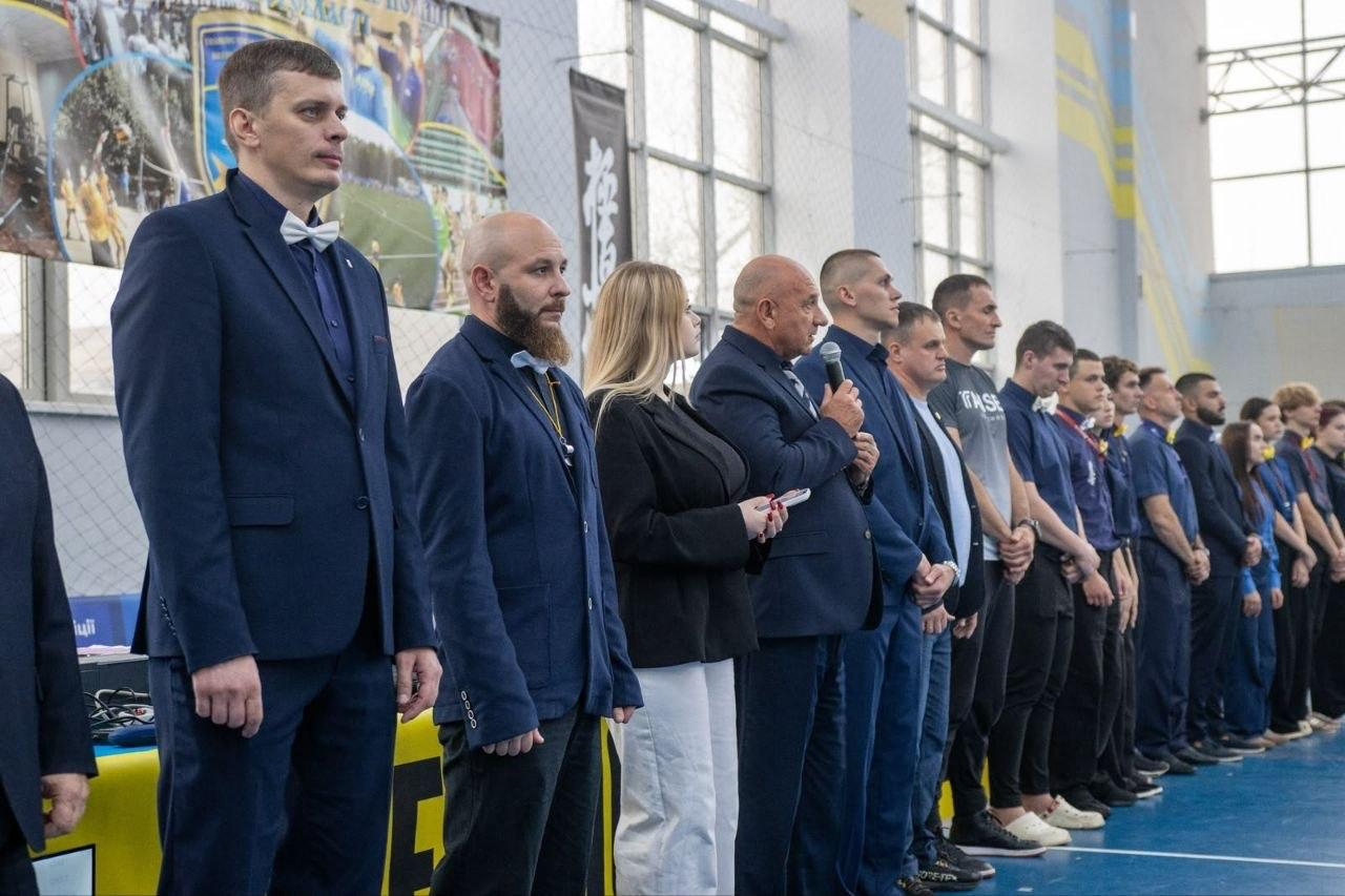 На світлині зафіксовано офіційну церемонію відкриття Чемпіонату України та Всеукраїнського турніру з кіокушин БуДо карате у Полтаві. У великому спортивному залі вздовж синього татамі в один шерег вишикувалися судді, почесні гості та організатори змагань. На передньому плані ліворуч стоїть високий суддя у темно-синьому класичному костюмі з білим метеликом, а поруч із ним — бородатий колега в окулярах та аналогічному строї з жовтим ланьярдом на шиї. По центру шеренги літній поважний чоловік у синьому піджаку тримає у руці мікрофон і виступає з вітальним словом перед учасниками. Поруч із ним стоять дівчина у чорному піджаку й білих штанях та інші представники суддівської колегії та тренери в офіційних костюмах, чорних футболках і спортивній формі, які урочисто слухають промову на тлі великих вікон спорткомплексу.