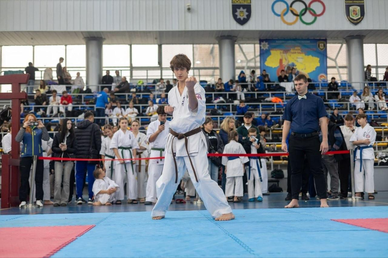 На світлині зафіксовано момент виступу юного спортсмена у розділі ката на Всеукраїнських змаганнях з кіокушин БуДо карате у Полтаві. На синьо-червоному покритті татамі по центру стоїть каратист у білому догі (кімоно) з коричневим поясом, який зосереджено виконує технічний комплекс, піднявши зігнуту в лікті ліву руку перед собою. Праворуч від нього уважно стежить за виконанням рефері на татамі у синій сорочці, чорних штанах та з білим метеликом. На задньому плані за червоною обмежувальною стрічкою зібралися глядачі та учасники змагань, а вище над заповненими трибунами стіну спорткомплексу прикрашають великі олімпійські кільця, емблеми організації та великий патріотичний банер із картою України.