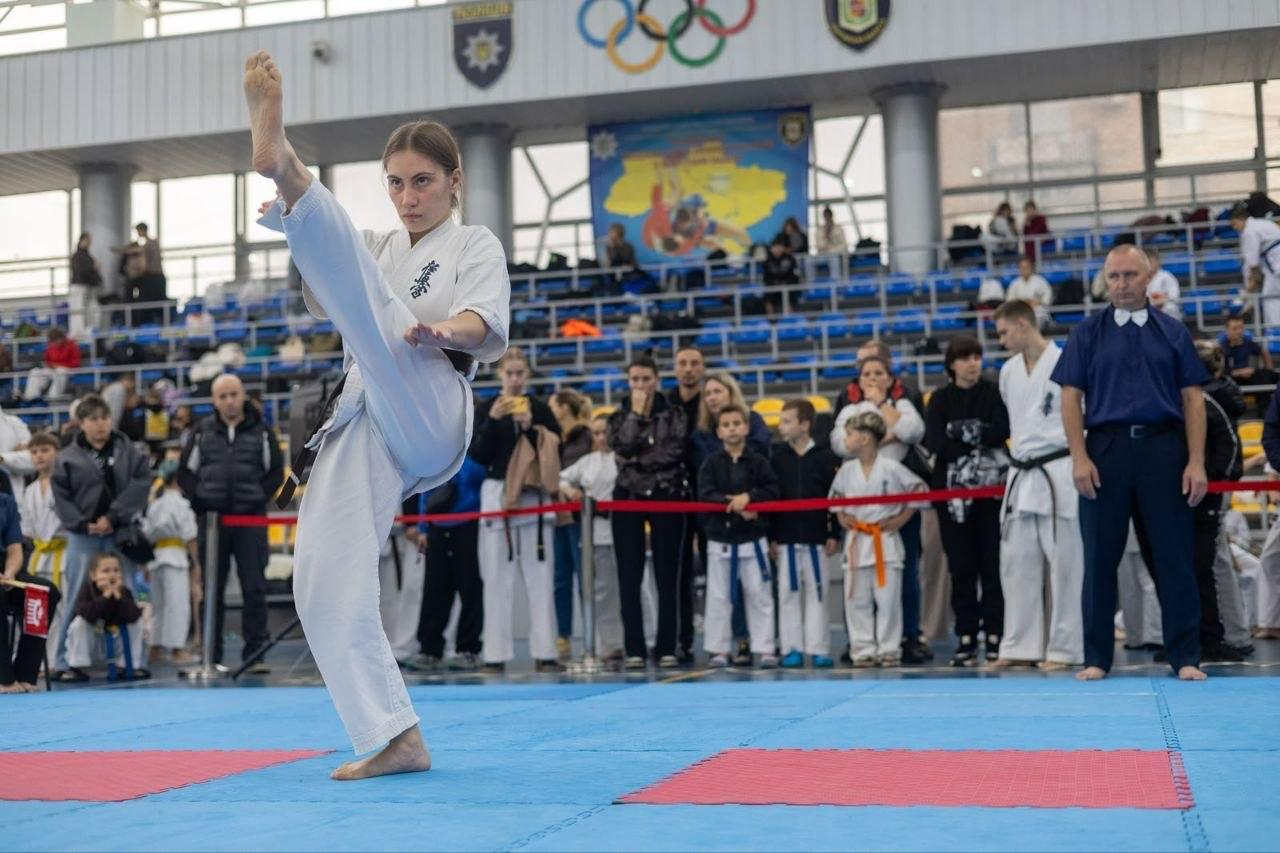 На світлині зафіксовано момент виступу спортсменки у розділі ката на Всеукраїнських змаганнях з кіокушин БуДо карате у Полтаві. Дівчина у білому кімоно з чорним поясом стоїть на одній нозі на синьо-червоному татамі, виконуючи високий удар ногою вгору (має-гері або йоко-гері) з максимальною концентрацією та тримаючи рівновагу. Праворуч від неї за виступом пильно спостерігає рефері у синій сорочці з білим метеликом. На задньому плані за червоною обмежувальною стрічкою зібралися глядачі, тренери та учасники турніру, а стіну спорткомплексу над трибунами прикрашають великі олімпійські кільця, емблеми та патріотичний банер із картою України.