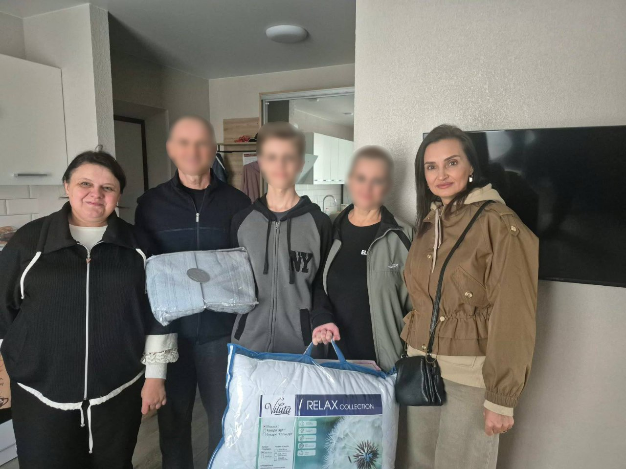 На світлині зафіксовано начальницю Олешківської МВА Тетяну Гасаненко під час візиту до евакуйованої родини з дітьми, яка змогла покинути тимчасово окуповану територію. У домашній кімнаті для спільного фото вишикувалися п'ятеро людей, при цьому обличчя трьох членів родини (двох дорослих та підлітка) на знімку штучно розмиті з міркувань безпеки, а чіткими залишаються обличчя представниць адміністрації. Крайня ліворуч стоїть представниця виконкому в чорній кофті на блискавці. Поруч із нею перебуває чоловік, який тримає в руках запакований комплект постільної білизни сірого кольору, а далі стоїть високий підліток у сіро-чорній кофті з написом «NY» та тримає велику синьо-білу фірмову сумку-упаковку з ковдрою або подушкою бренду «Viluta | RELAX COLLECTION». Праворуч від підлітка стоїть жінка в сірій кофті, а крайня праворуч — очільниця МВА Тетяна Гасаненко у коричневій куртці з капюшоном та чорною сумочкою через плече. Зустріч проходить у квартирі зі світлими стінами, де на задньому плані видно великий плазмовий телевізор та дзеркальну шафу-купе.