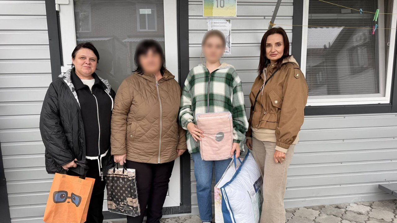 На світлині зафіксовано начальницю Олешківської МВА Тетяну Гасаненко під час візиту до ще однієї евакуйованої родини з Олешок. Біля входу до модульного або тимчасового будинку зі світло-сірою сайдинговою обшивкою стін для спільного фото вишикувалися четверо людей, при цьому обличчя жінки та дівчини-підлітка штучно розмиті з міркувань безпеки. Крайня ліворуч стоїть представниця виконкому в чорній куртці та чорній кофті, яка тримає в руці помаранчевий подарунковий пакет із малюнком сови в окулярах. Поруч із нею стоїть евакуйована жінка в коричневій стьобаній куртці з темним пакетом у руці, а далі — дівчина-підліток у кардигані в зелено-білу клітинку та синіх джинсах, яка тримає запакований рожевий комплект постільної білизни та велику фірмову сумку-упаковку «Viluta | RELAX COLLECTION». Крайня праворуч позує очільниця МВА Тетяна Гасаненко у коричневій куртці з капюшоном та світло-бежевих штанях. Над головами присутніх на стіні будинку помітно табличку з номером «10» та невелике інформаційне оголошення.