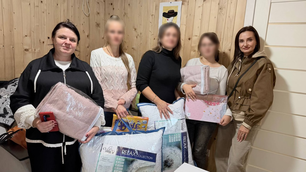 На світлині зафіксовано начальницю Олешківської МВА Тетяну Гасаненко під час візиту до третьої евакуйованої родини з Олешок. У кімнаті з оздобленими світлим деревом стінами для спільного фото вишикувалися п'ятеро жінок та дівчат, при цьому обличчя трьох евакуйованих мешканок у центрі кадру штучно розмиті з міркувань безпеки. Крайня ліворуч стоїть представниця виконкому в чорній кофті на блискавці, яка тримає в одній руці смартфон, а в іншій — великий запакований рожевий рушник або ковдру. Поруч із нею дівчина у світлому светрі з візерунками тримає коробку з настільною грою «Monopoly». По центру жінка в чорному гольфі тримає велику синьо-білу фірмову сумку-упаковку «Viluta | RELAX COLLECTION», а праворуч від неї дівчина у світлій кофті тримає запакований комплект постільної білизни та ще одну подушку. Крайня праворуч позує очільниця МВА Тетяна Гасаненко у коричневій куртці з капюшоном та світло-бежевих штанях. На дерев'яній стіні позаду присутніх помітно невеликий малюнок або постер із зображенням жінки в капелюсі.