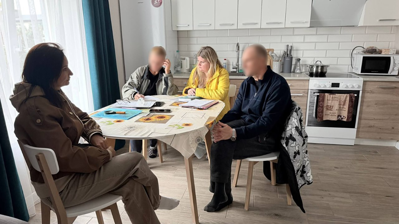 На світлині зафіксовано процес надання соціальних і правових консультацій евакуйованій родині з Олешок начальницею Олешківської МВА Тетяною Гасаненко. За обіднім столом із квітковою скатертиною на сучасній світлій кухні сидять четверо людей, при цьому обличчя евакуйованого чоловіка праворуч та хлопчика-підлітка на задньому плані штучно розмиті з міркувань безпеки. Ліворуч у профілі на передньому плані на дерев'яному стільці сиділа очільниця МВА Тетяна Гасаненко у коричневій куртці з капюшоном та світлих штанях, яка уважно слухає співрозмовників. Навпроти неї сидить чоловік у темно-синій кофті, а поруч із ним за столом перебуває представниця виконкому в яскравій жовтій куртці, яка розбирає документи та робить записи в журналі. На столі перед ними розкладено папери, лежить лупа, окуляри та смартфони. На задньому плані кадру детально видно інтер'єр кухні: білий холодильник, світлі кухонні шафки, білу кахельну плитку «кабанчик» на фартуху, робочу поверхню з кухонним приладдям, каструлею на плиті та мікрохвильову піч праворуч.