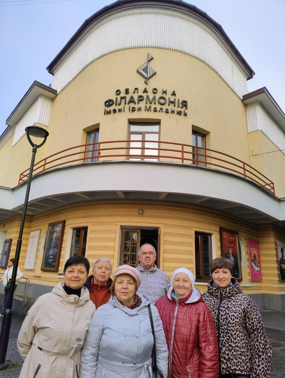 На світлині зафіксовано групу внутрішньо переміщених осіб із Херсонщини на тлі головного фасаду Івано-Франківської обласної філармонії імені Іри Маланюк, візит до якої організував хаб «Вільні разом». Шість відвідувачів вишикувалися на тротуарі перед будівлею зліва направо: на передньому плані ліворуч стоїть жінка у світло-кремовому пальті, трохи позаду неї — жінка у помаранчевій стьобаній куртці, по центру попереду позує жінка у довгій світло-сірій куртці та світло-рожевому капелюшку, за її спиною стоїть літній сивочолий чоловік у сірій дутій куртці, правіше стоїть жінка у бордовому пуховику з капюшоном, а крайня праворуч — жінка у куртці з леопардовим принтом. На задньому плані височіє жовто-біла напівкругла архітектурна вежа будівлі філармонії з балюстрадою балкона, де великими чорними літерами нанесено напис «ОБЛАСНА ФІЛАРМОНІЯ імені Іри Маланюк» та емблема, а на стінах біля вікон першого поверху розвішані афіші концертів, зокрема рожевий плакат «Музика українського кіно».