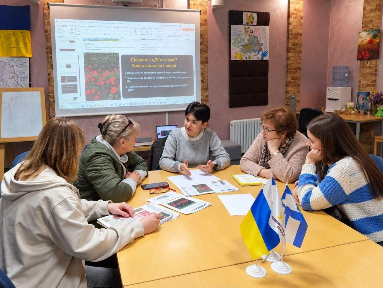 На світлині зафіксовано проведення інформаційної акції «Втрачені документи на землю: алгоритм дій» для ВПО з Херсонщини в ужгородському хабі «Вільні разом». За великим столом сидять п'ять жінок, де в центрі лекторка з короткою зачіскою у сірому светрі роз'яснює юридичні алгоритми слухачкам, які вивчають друковані брошури. На передньому плані встановлено настільні прапорці Херсонської області та Нижньосірогозької СВА, а на задньому плані на великий екран виведено слайд презентації з інструкцією: «Ділянки в «Дії» немає? Права лише на папері» та покроковим алгоритмом дій щодо реєстрації прав на нерухомість.