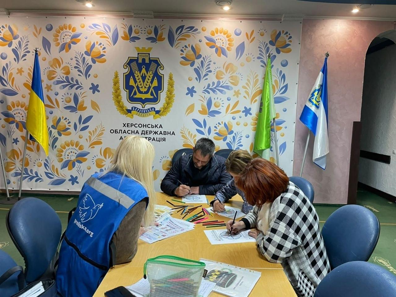 На світлині зафіксовано групу психологічної підтримки «Пошук ресурсу» для внутрішньо переміщених осіб із Херсонщини, яку в ужгородському хабі «Вільні разом» провела фахівчиня Гуманітарної місії «Проліска». За великим овальним столом світлого дерева сидять чотири людини, які займаються арттерапевтичним малюванням: на передньому плані ліворуч спиною до об'єктива сидить психолог у синьому фірмовому жилеті з емблемою білого птаха та адресою сайту proliska.org, а навпроти неї перебувають три учасники заходу (чоловік у темній куртці, жінка у синій куртці з візерунками та жінка в картатій чорно-білій кофті), які зосереджено розфарбовують круглі мандали-антистрес. У центрі столу розкладено велику кількість різнокольорових олівців, лежать друковані методичні матеріали, а на передньому плані стоїть прозора зелена сумочка-пенал з канцелярією. Зустріч проходить у залі на тлі великого офіційного білого банера, який прикрашений яскравими жовто-блакитними рослинними орнаментами петриківського розпису, золотистим гербом Херсонської області та великим чорним написом «ХЕРСОНСЬКА ОБЛАСНА ДЕРЖАВНА АДМІНІСТРАЦІЯ». По боках від банера встановлено Державний Прапор України, а праворуч — прапор Херсонської області та світло-зелений прапор.