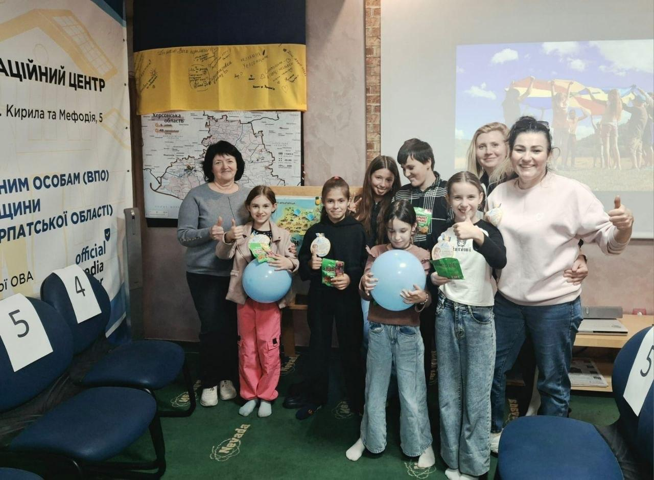 На світлині в центрі кадру позує група дітей різного віку: ліворуч на передньому плані стоїть дівчинка у світлій куртці, яскраво-рожевих спортивних штанях та білих кросівках, яка тримає велику блакитну повітряну кульку та пакуночок із солодощами; поруч із нею стоїть дівчинка в чорному худі з капюшоном, чорних штанях та темних кросівках, яка також тримає в руці святковий сувенір; правіше перебуває дівчинка у блакитних джинсах, коричневій кофті та світлих шкарпетках, яка двома руками утримує перед собою ще одну велику блакитну надувну кулю; біля неї праворуч радісно посміхається дівчинка у світло-блакитних джинсах та білому світшоті з патріотичним малюнком і написом, яка показує жест «клас» піднятим догори великим пальцем; трохи позаду них по центру стоять двоє підлітків — дівчина з довгим русявим волоссям у чорній кофті та хлопчик у картатій зелено-чорній сорочці поверх темного худі, який тримає в руках солодкий пакунок. Разом із дітьми на знімку присутні жінки-організатори та матусі: крайня ліворуч за спинами дівчаток стоїть жінка старшого віку у сірому джемпері, яка посміхається і піднімає великий палець вгору; у правій частині кадру позаду дітей стоїть жінка у чорній кофті, яка ніжно обіймає підлітків за плечі, а попереду неї радісно позирує ще одна жінка у світлому кремовому светрі та синіх джинсах, яка впевнено показує жест «клас» обома руками.  На задньому плані ліворуч видно інформаційний стенд координаційного центру, карту Херсонської області та підписаний патріотичними побажаннями Державний Прапор України, а праворуч на великий мультимедійний екран виведено слайд із яскравою літньою фотографією дітей біля річки під синім небом.