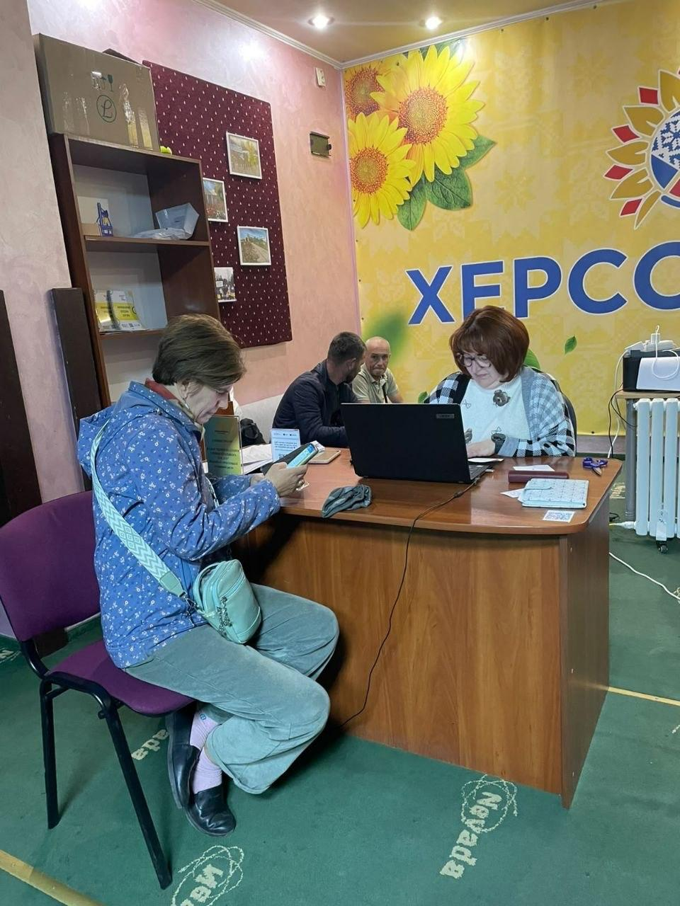 На світлині зафіксовано надання правових консультацій для внутрішньо переміщених осіб із Херсонщини в ужгородському хабі «Вільні разом». На передньому плані ліворуч на фіолетовому стільці сидить жінка в синій куртці в дрібну квіточку та сірих вельветових штанях із ніжно-зеленою сумочкою через плече, яка зосереджено дивиться у свій смартфон. Навпроти неї за великим кутовим дерев'яним столом сидить фахівчиня в окулярах та картатій чорно-білій кофті, яка перевіряє інформацію на відкритому чорному ноутбуці. Два чоловіки (один у темній куртці, інший — у світлій сорочці) зафіксовані трохи позаду на дивані, очікуючи на свою чергу або відпочиваючи під час прийому. Подія розгортається в робочому кабінеті на тлі великого офіційного яскраво-жовтого банера з малюнком соняшників та початком синього напису «ХЕРСОН», а ліворуч на стіні помітно дерев'яний стелаж із брошурами та бордовий стенд із фотокартками краєвидів.