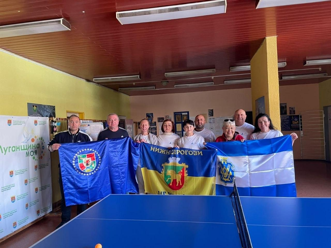 На світлині зафіксовано учасників та організаторів дружнього турніру з настільного тенісу «Вільні разом — спортивні разом», який об'єднав внутрішньо переміщених осіб із Херсонщини та Луганщини в ужгородському координаційному центрі «Вільні разом». На передньому плані встановлено синій тенісний стіл із натягнутою сіткою, за яким для спільного фото вишикувалася група з дев'яти людей (чотирьох чоловіків та п'яти жінок), які з посмішками тримають три великі прапори. Ліворуч двоє чоловіків у темних спортивних кофтах тримають синє знамено Луганської області з її офіційним гербом, по центру група жінок та чоловік у білих фірмових футболках тримають синьо-жовтий прапор Нижньосірогозької селищної ради з написом «НИЖНІ СІРОГОЗИ» та зображенням золотого лева на червоному щиті, а праворуч представники тримають синьо-білий прапор Херсонської області. Крайнім ліворуч у залі помітно білий банер із патріотичним написом «Луганщина моя», стіни приміщення пофарбовані у світло-жовтий колір, а коричнева дерев'яна стеля облаштована довгими люмінесцентними лампами.