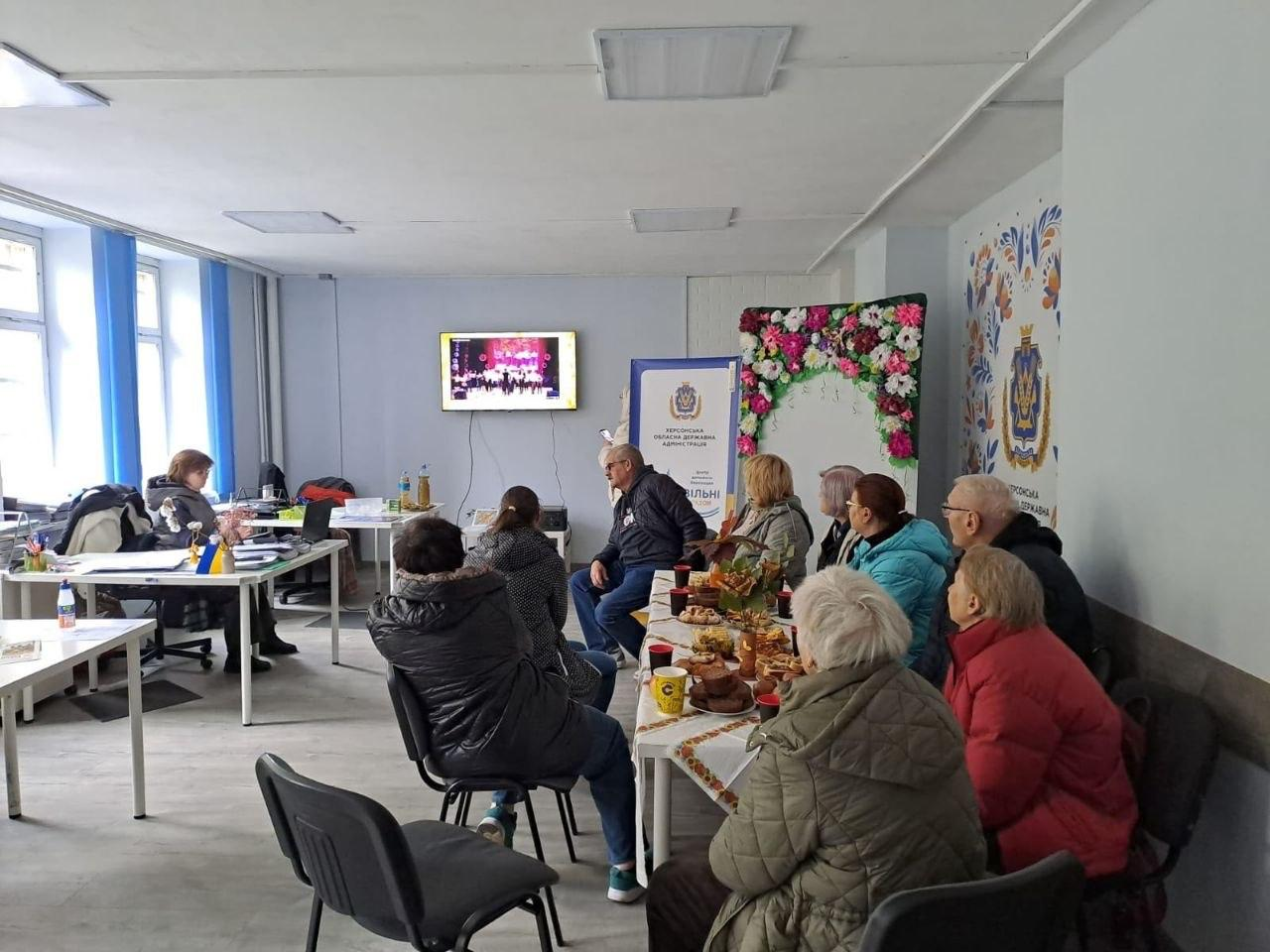 На світлині зафіксовано зустріч внутрішньо переміщених осіб із Херсонщини у координаційному центрі «Вільні разом» у Львові, де відвідувачі переглядають відеоматеріал «Херсонщина культурна» та відпочивають біля тематичної фотозони. На передньому плані та по центру навколо довгого столу, накритого білою скатертиною з українським вишитим орнаментом, сидить група з дев'яти літніх людей у верхньому осінньому одязі, які повернуті обличчям до дальньої стіни; на столі перед ними розставлено різнокольорові кухлі, тарілки з печивом та осінню композицію з жовтого листя. На дальній сірій стіні по центру закріплено плазмовий телевізор, на екрані якого транслюється концертна програма з артистами на сцені. Праворуч на стіні розташована ошатна квіткова арка-фотозона, густо прикрашена рожевими, білими та бузковими квітами з зеленим листям, яка знаходиться між мобільним білим ролап-банером з лівого боку, а з правого боку на білій стіні нанесено герб Херсонської області з яскравим рослинним орнаментом петриківського розпису та написом «ХЕРСОНСЬКА ОБЛАСНА ДЕРЖАВНА АДМІНІСТРАЦІЯ». Ліворуч у кімнаті біля великих вікон із довгими блакитними шторами розміщено офісні столи, де за ноутбуком та документами сидить фахівчиня центру у сірому светрі, яка спілкується з відвідувачкою.