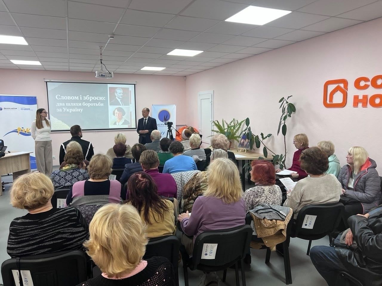На світлині показано засідання розмовно-інтеграційного клубу «РІКА» для переселенок із Херсонщини, що тимчасово проживають у Кропивницькому. У просторому світлому залі з ніжно-рожевими стінами зібралася велика аудиторія слухачок, які сидять на чорних стільцях та слухають лекторів біля великого проекційного екрана. На екрані продемонстровано слайд презентації з портретами Романа Шухевича та Олени Теліги й великим написом: «Словом і зброєю: два шляхи боротьби за Україну». Ліворуч перед слухачами стоїть молода лекторка у світлому вбранні, а по центру біля стіни виступає чоловік у класичному темному костюмі та краватці, поруч із яким розташовані мобільні банери хабу «РІКА» та ГО «Десяте квітня». Справа на стіні встановлено великий об'ємний помаранчевий логотип гуманітарного центру, а на передньому плані кадру видно відвідувачок у теплих осінніх кофтах та куртках, які тримають у руках друковані інформаційні брошури.