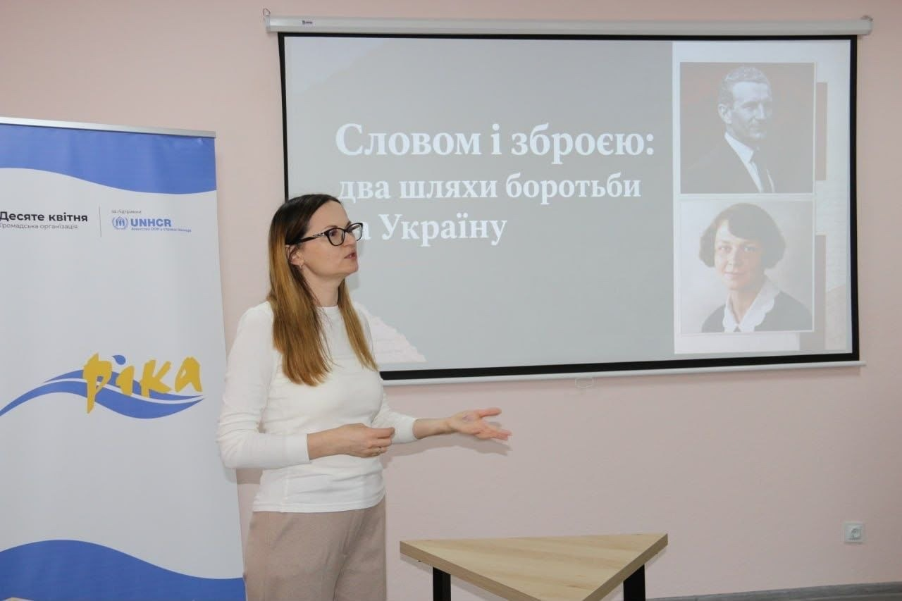 На світлині показано молоду лекторку розмовно-інтеграційного клубу «РІКА» під час виступу на заході «Словом і зброєю: два шляхи боротьби за Україну» в Кропивницькому. Дівчина в окулярах, одягнена у світлий білий лонгслів та бежеві штани, стоїть у центрі кадру біля великого проекційного екрана й активно жестикулює руками, роз'яснюючи матеріал слухачам. На екрані позаду неї демонструється слайд презентації з великим білим заголовком заходу, а праворуч наведено чорно-білі портрети видатних діячів українського національно-визвольного руху — Романа Шухевича та Олени Теліги. Ліворуч від доповідачки встановлено біло-блакитний мобільний ролап-банер із жовтим художнім написом «РІКА», логотипами громадської організації «Десяте квітня» та Агентства ООН у справах біженців «UNHCR», а на передньому плані перед екраном помітно частину світлого дерев'яного столу.