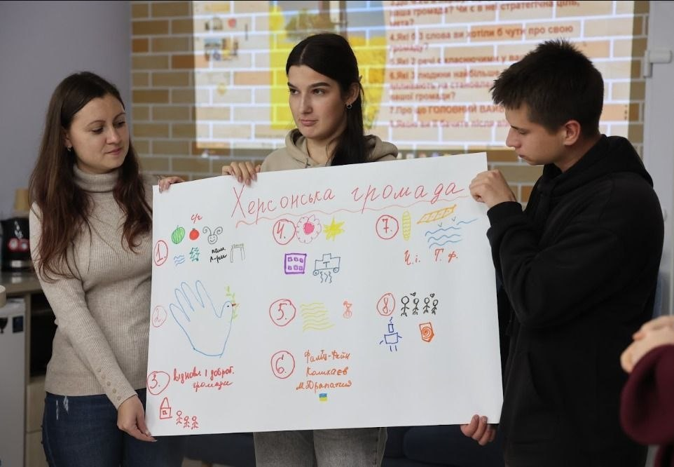 На світлині показано презентацію напрацювань учасників тренінгу з локального брендування в Миколаєві, які представляють заклади культури та освіти Херсонщини. Троє молодих людей (дві дівчини та юнак) спільно тримають великий білий ватман із проєктом під назвою «Херсонська громада», прикрашеним кольоровими малюнками, символами та текстовими нотатками за номерами. Ліворуч стоїть дівчина у світло-бежевому гольфі та синіх джинсах, по центру — дівчина у світло-коричневому худі, а праворуч — юнак у чорній кофті з капюшоном, який зосереджено дивиться на плакат. На ватмані серед малюнків помітно контури долоні з голубем миру, кавуна, помідора, колосків, хвиль Республіки та підпис «відбудова і добробут громади». На задньому плані кадру на світло-коричневу цегляну стіну виведено слайд презентації з переліком запитань, серед яких чітко видно пункти: «Які 3 слова ви хотіли б чути про свою громаду?» та «Які 3 речі є класичними у вашій громаді?».