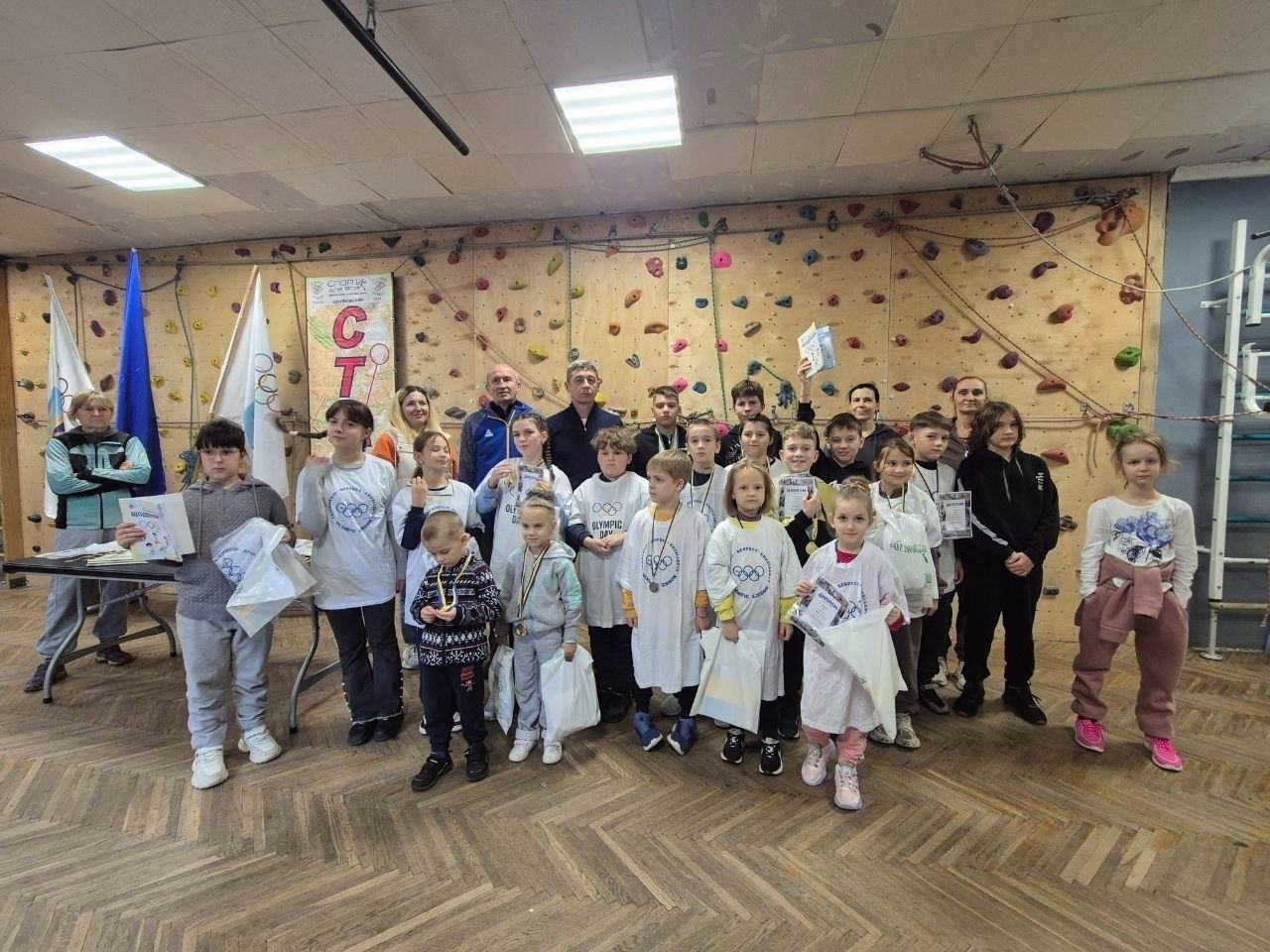 На світлині показано велику групу юних спортсменів, тренерів та організаторів мінітурніру зі спортивного туризму на Херсонщині, присвяченого четвертій річниці визволення правобережжя області від російських окупантів. У центрі спортивної зали на тлі дерев'яної стіни-скеледрому з різнокольоровими зачепами для скелелазіння вишикувалися близько 30 учасників віком від 6 до 14 років, які радісно позують із завойованими дипломами, медалями на шиї та білими пакунками з призами. Багато дітей одягнені у фірмові білі футболки з п'ятьма олімпійськими кільцями, емблемою та синіми патріотичними написами. У задньому ряду по центру стоять дорослі тренери та судді, серед яких чоловік у синій спортивній кофті та чоловік у чорній куртці, ліворуч біля столу з нагородами перебуває жінка-організатор у бірюзовому жилеті, а поруч майорять Державний Прапор України та білі прапори з олімпійською символікою.